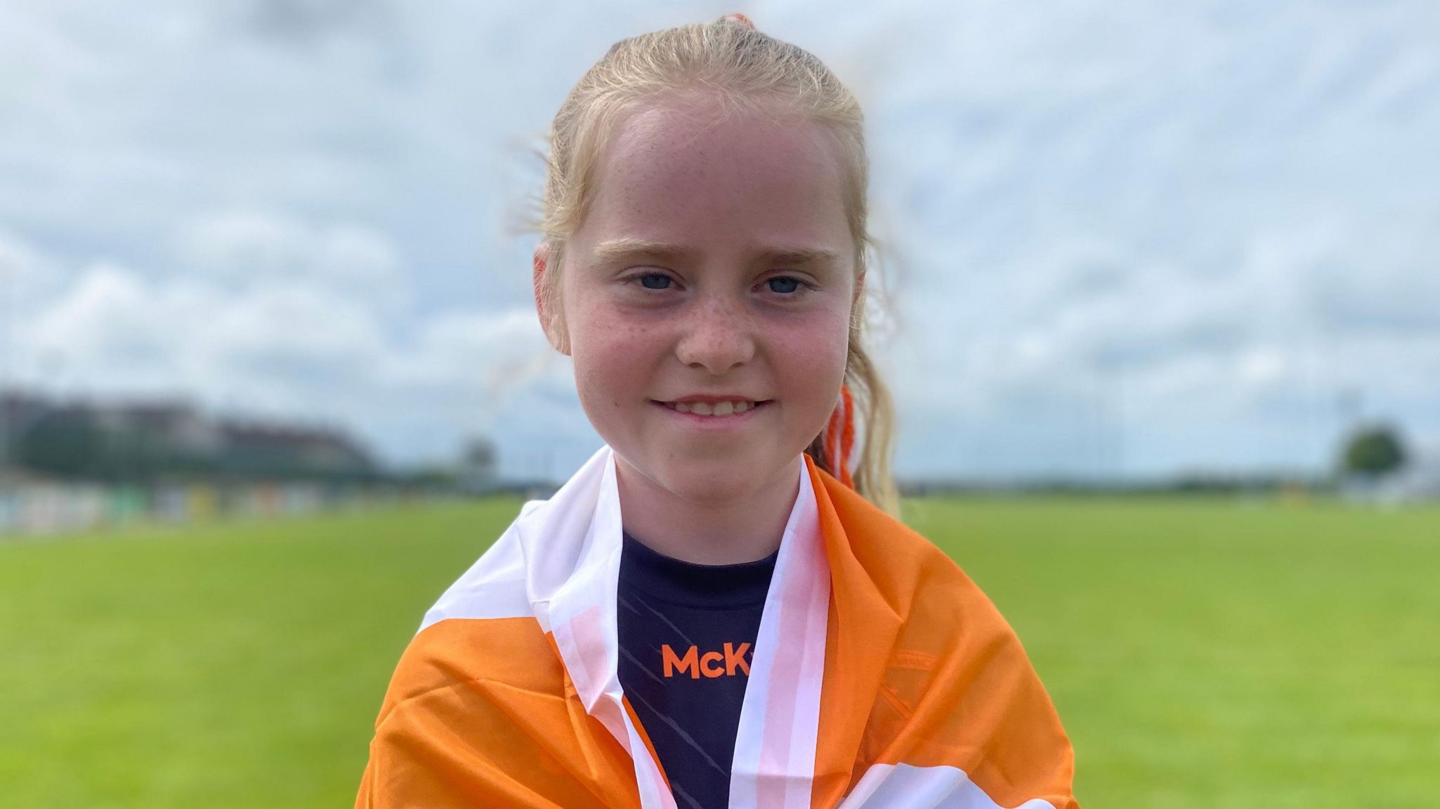 Sarah is smiling into the camera. she has her blonde hair tied into a ponytail and has an orange and white flag draped around her shoulders.