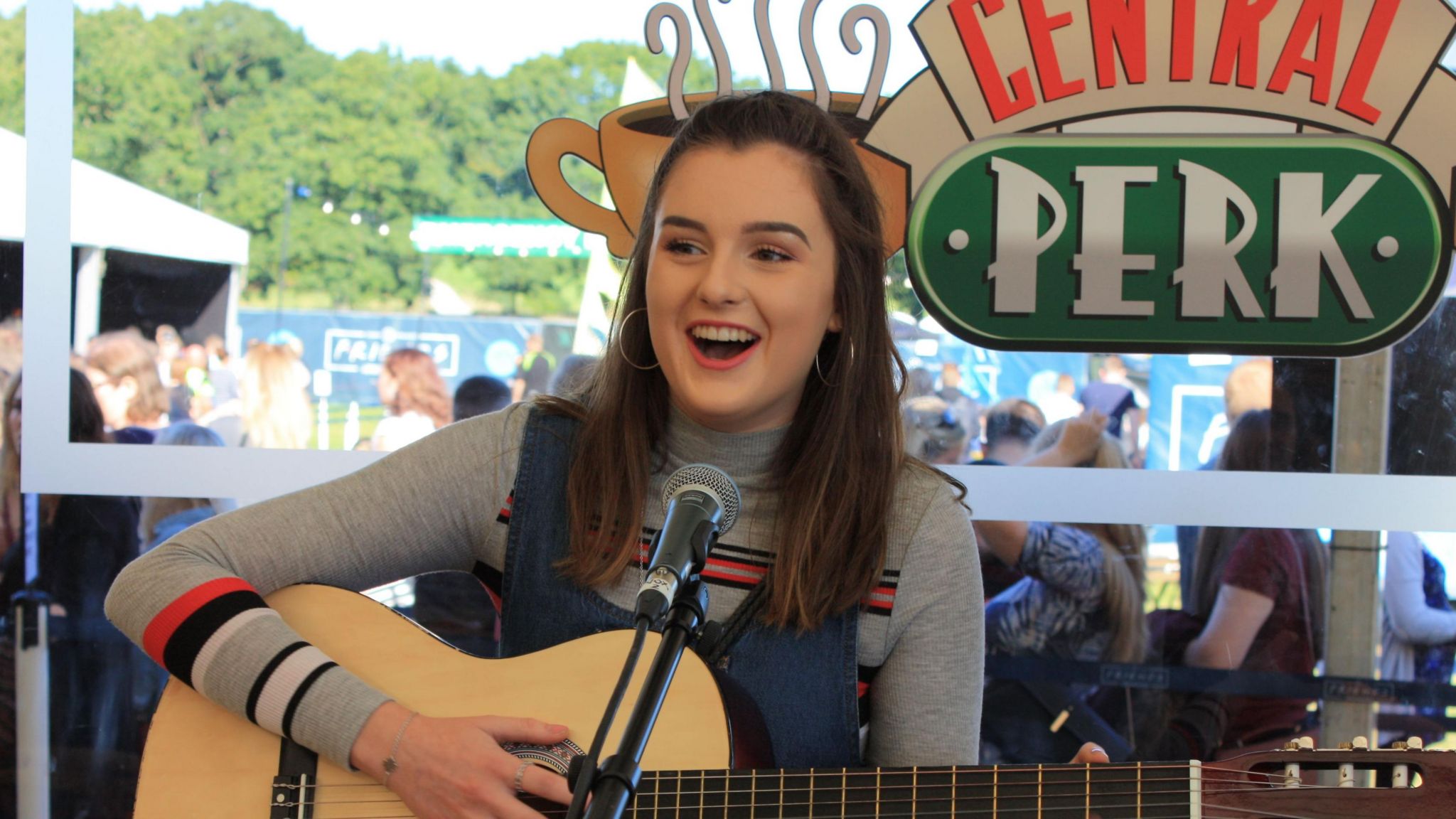 Beth Langton, who has long brown hair, looks off to the left as she poses for a photo. She is holding a guitar and there is a microphone in front of her