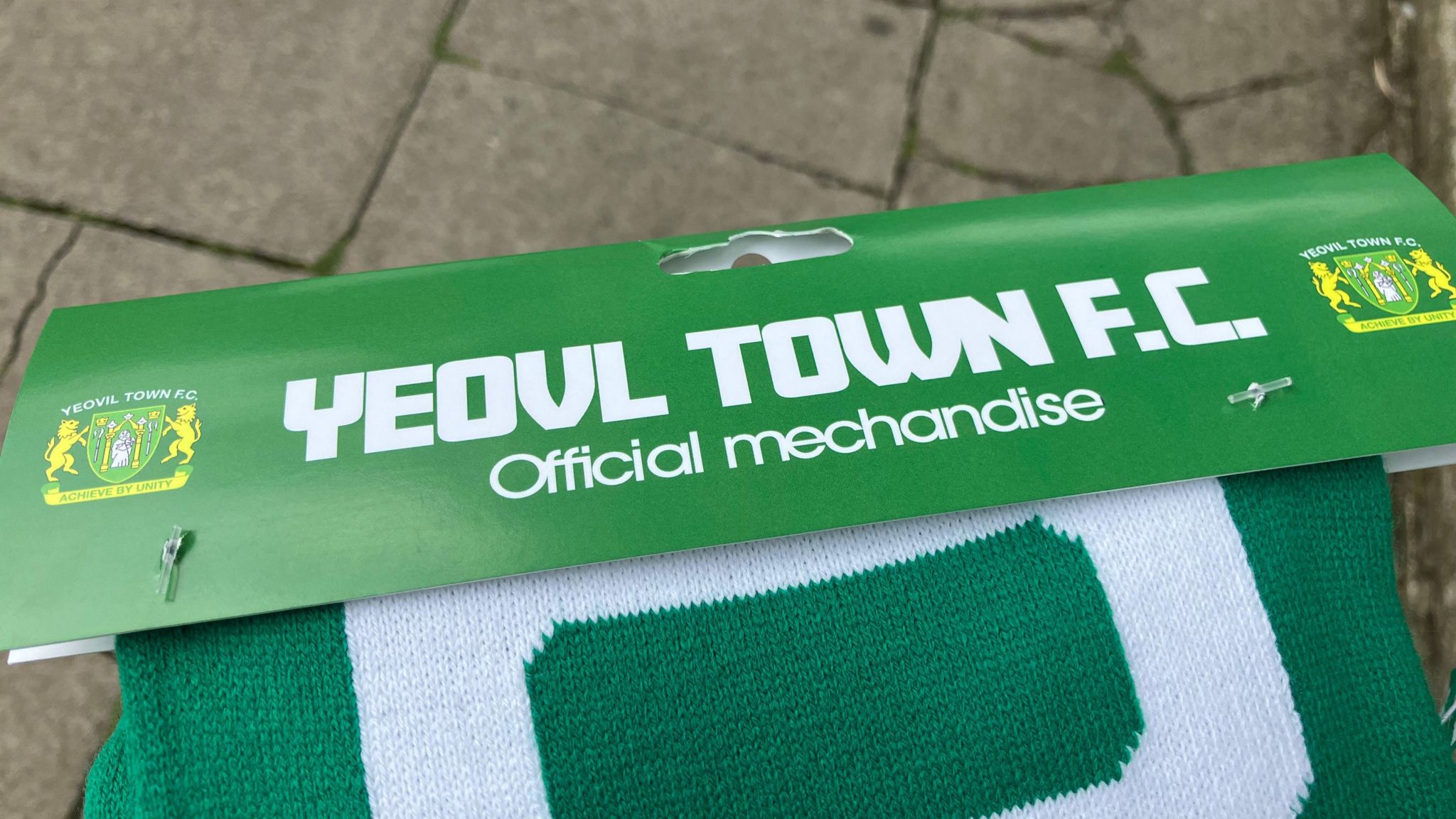 Picture of green Yeovil scarf with misspellings on the packaging including "Yeovl" without the i, and has misspelt the word merchandise without an "r".