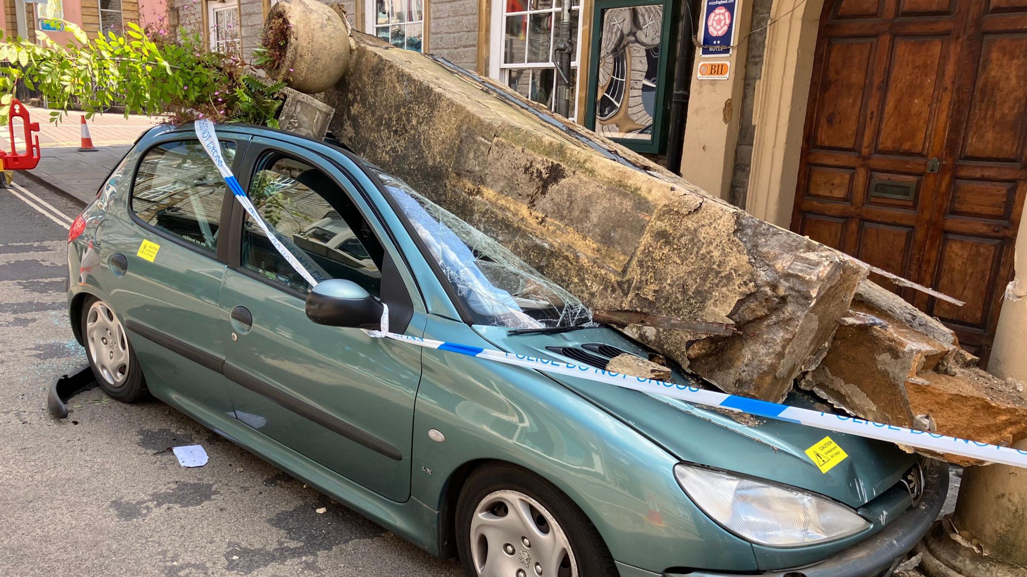 Image of a green Peugeot which crashed. Police tape can be seen around the vehicle, and a large pillar is on top of the windscreen.