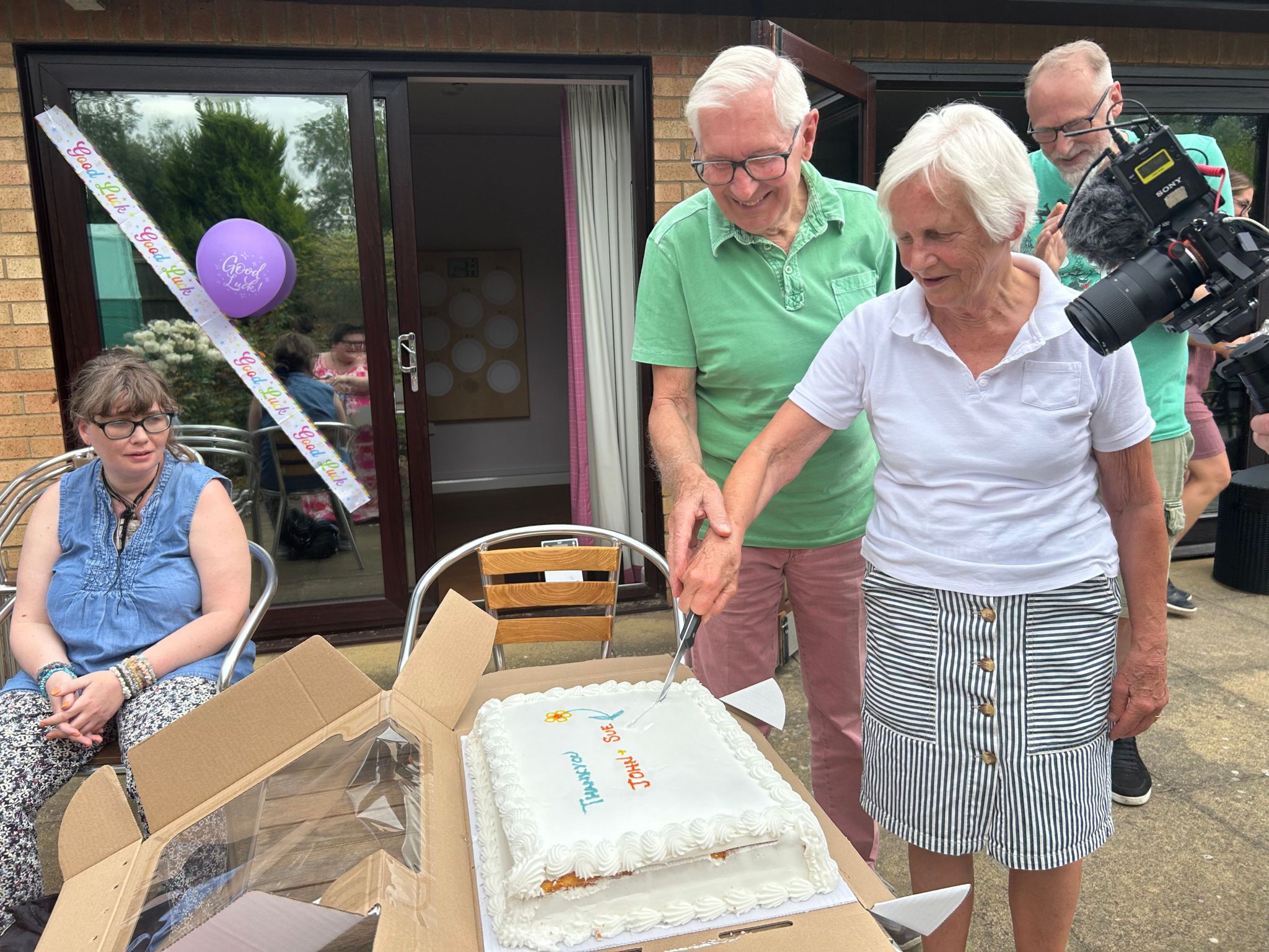 A man in a green polo and a woman in a white polo cut a cake while other people look on 