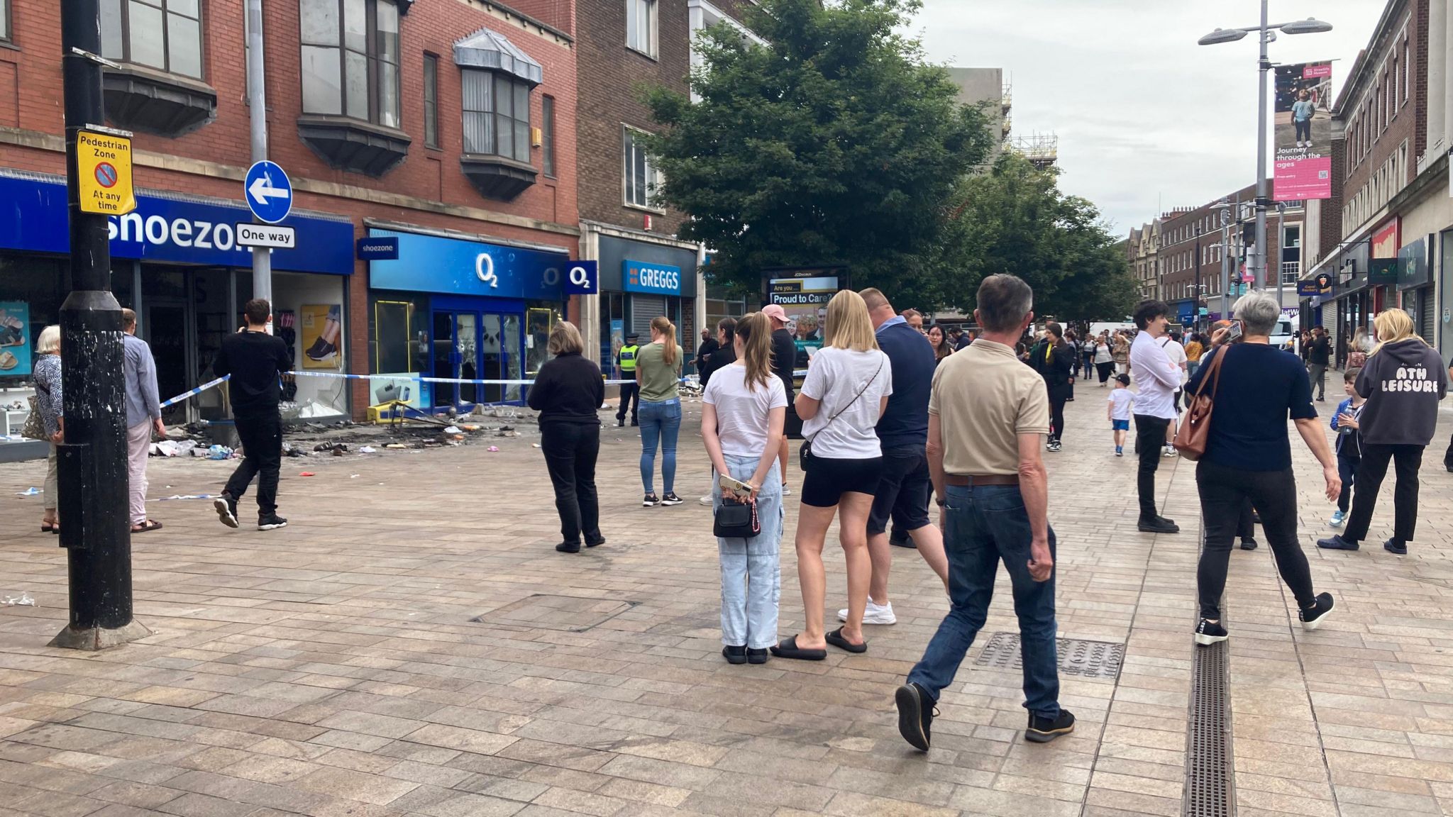 A man wearing blue jeans and a cream T-shirt walks by as other members of the public stand and look at damage on the street