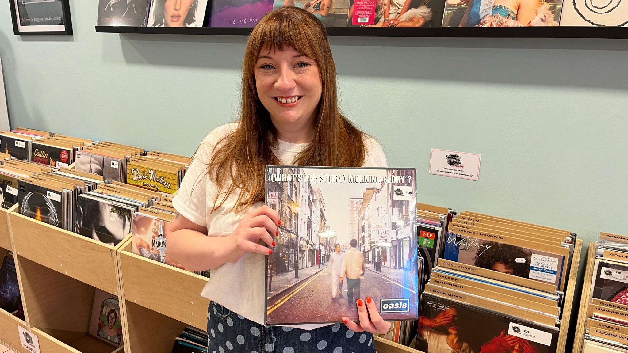 A woman with brown hair holds a vinyl copy of Oasis album What's the Story Morning Glory with a row of records behind her.