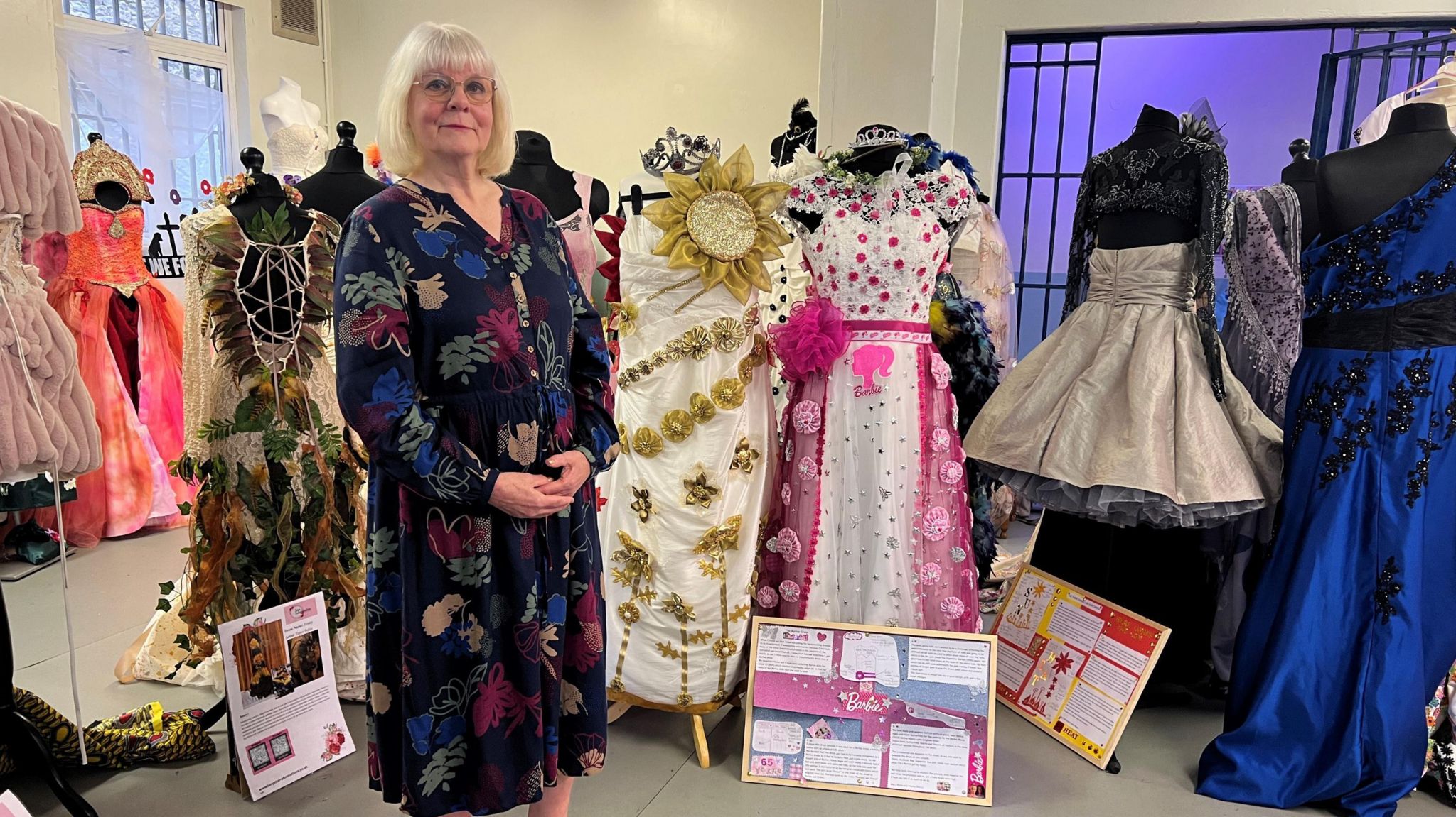 Marjorie Wells standing next to her dresses - one is white with gold flowers wrapped around it and a sunflower at the top. The other is white with pink flowers decorating it.