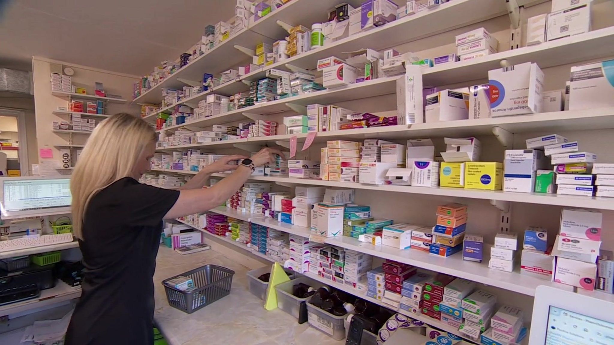 A woman with blonde hair and wearing a black medical uniform taking prescription drug boxes from one of the fifth row of shelves