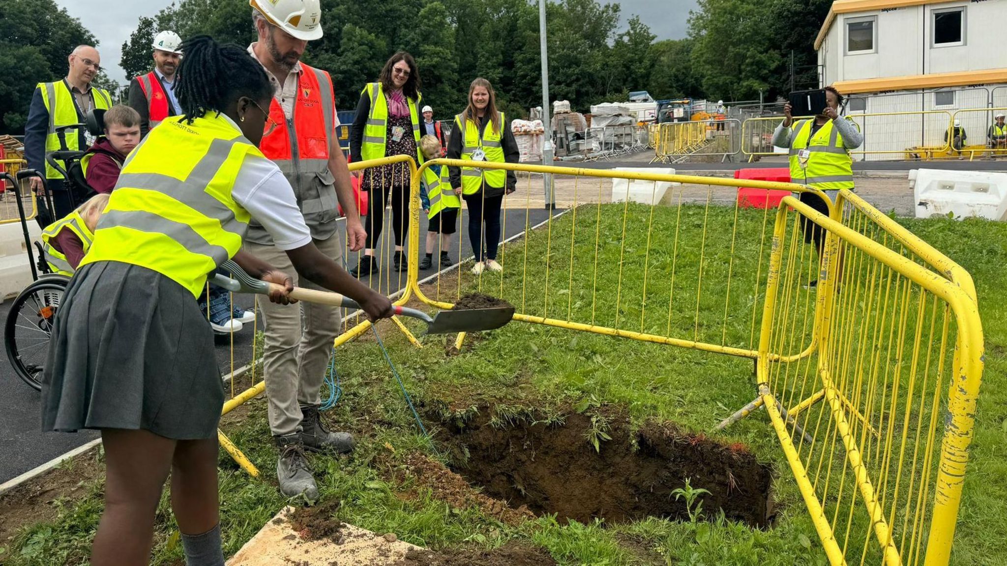 A school girl wearing a high-vis jacket and school skirt, shovelling soil into a hole to cover a time capsule on a field outside the school, with other teachers and people looking on.