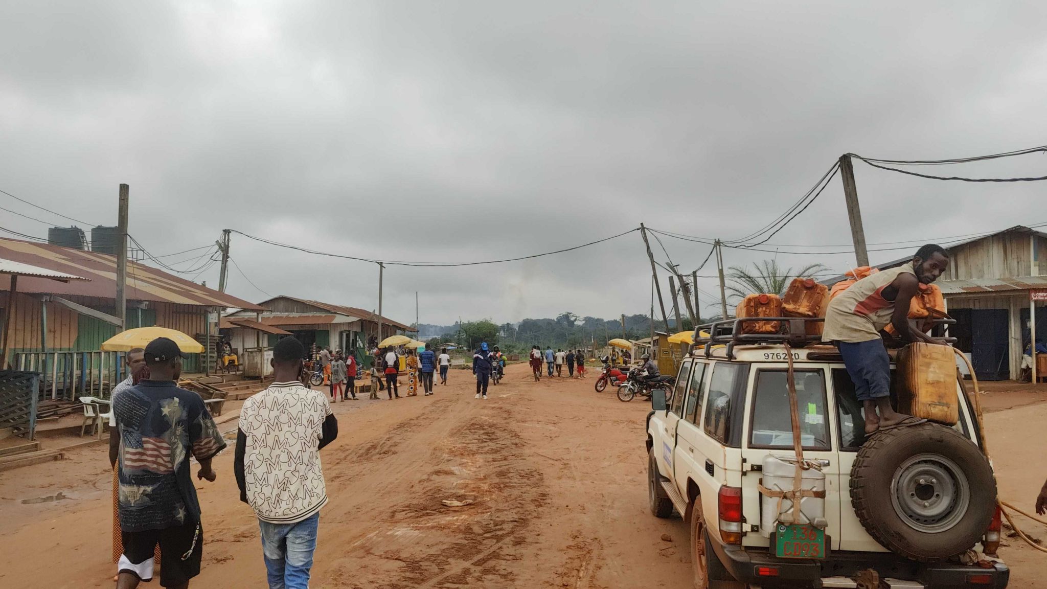 A dirt road in the Republic of Congo. People can be seen walking in the road, and a man on a 4x4 vehicle can be seen standing on a spare tyre attached to the back of it.