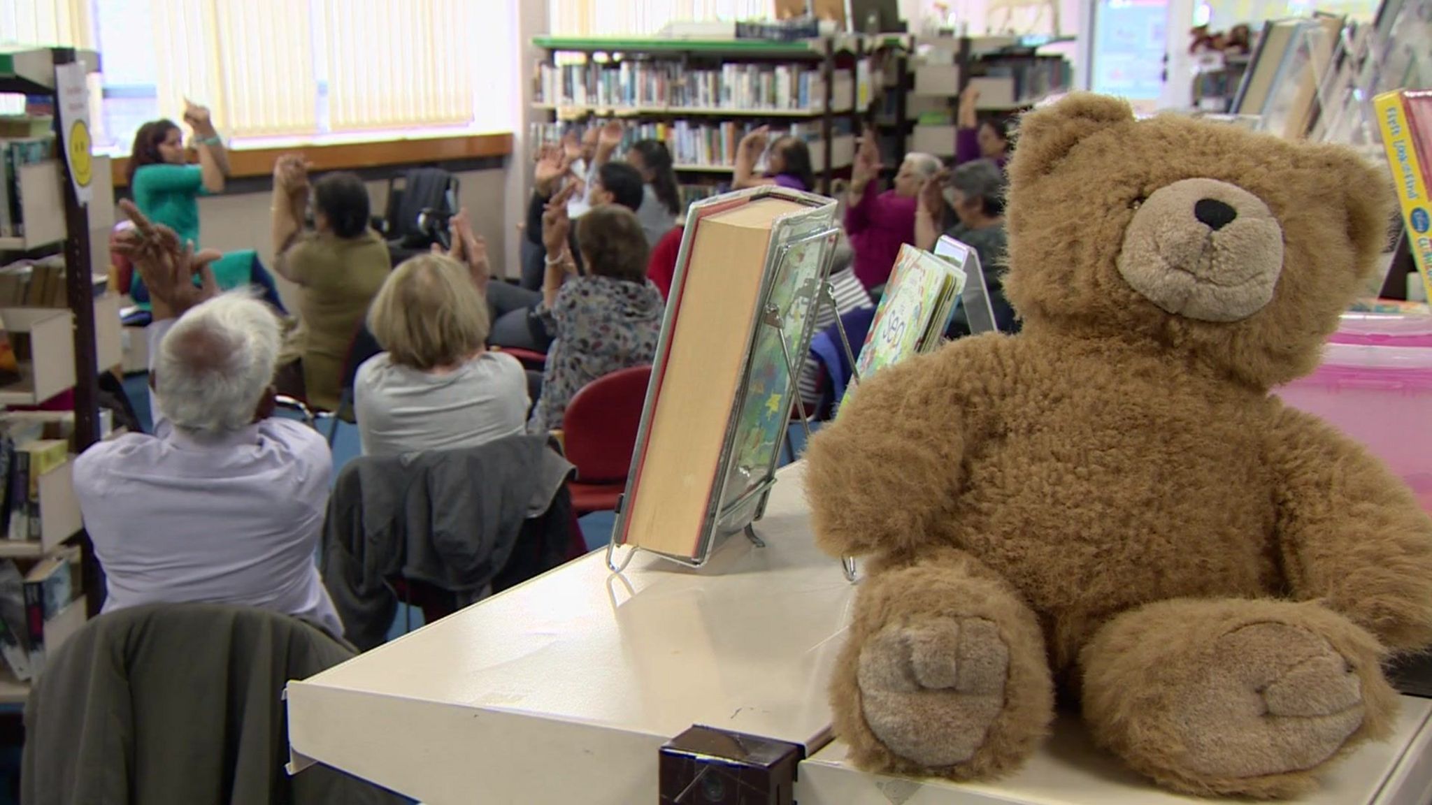 A group of people with their backs to the camera do chair yoga in the library.