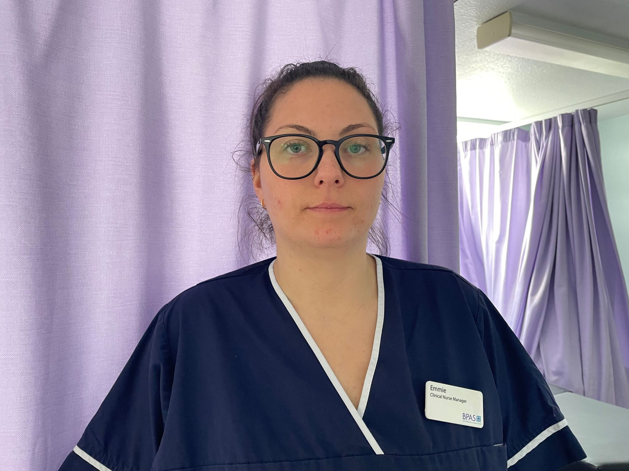 Emmie Isaac wearing a navy blue nurse's uniform and badge in a hospital ward with purple cutains. She is wearing black-rimmed, round glasses and her hair is tied back