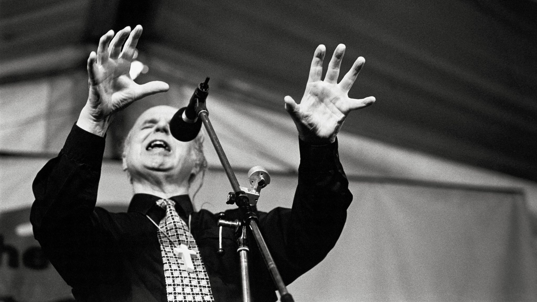 Black and white image of the the leader of the Jesus Fellowship, Noel Stanton, giving a sermon with his arms raised in front of him