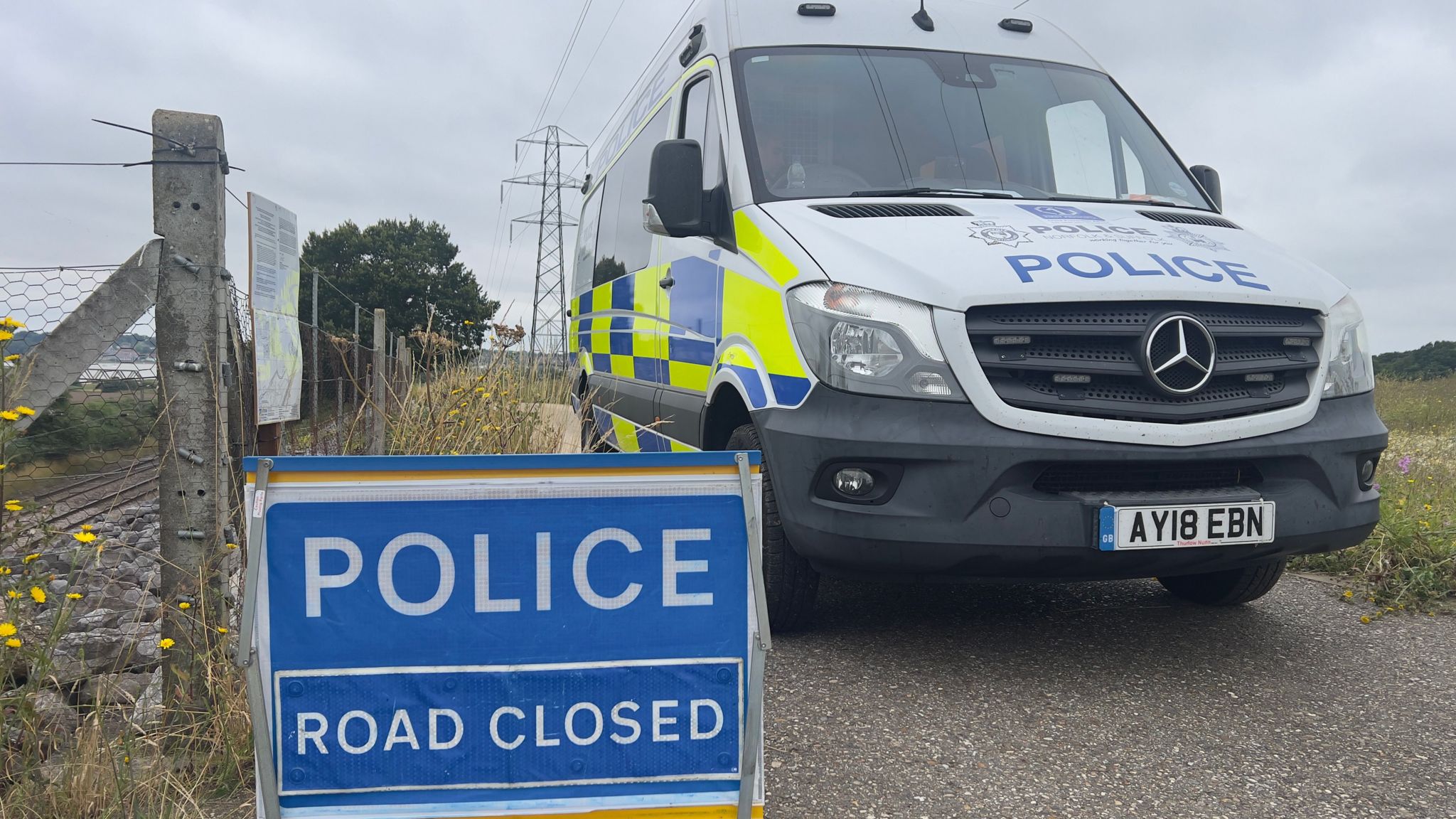 A police van and a sign that reads "Police Road Closed", both of which are blocking the entrance to a lane by a railway track