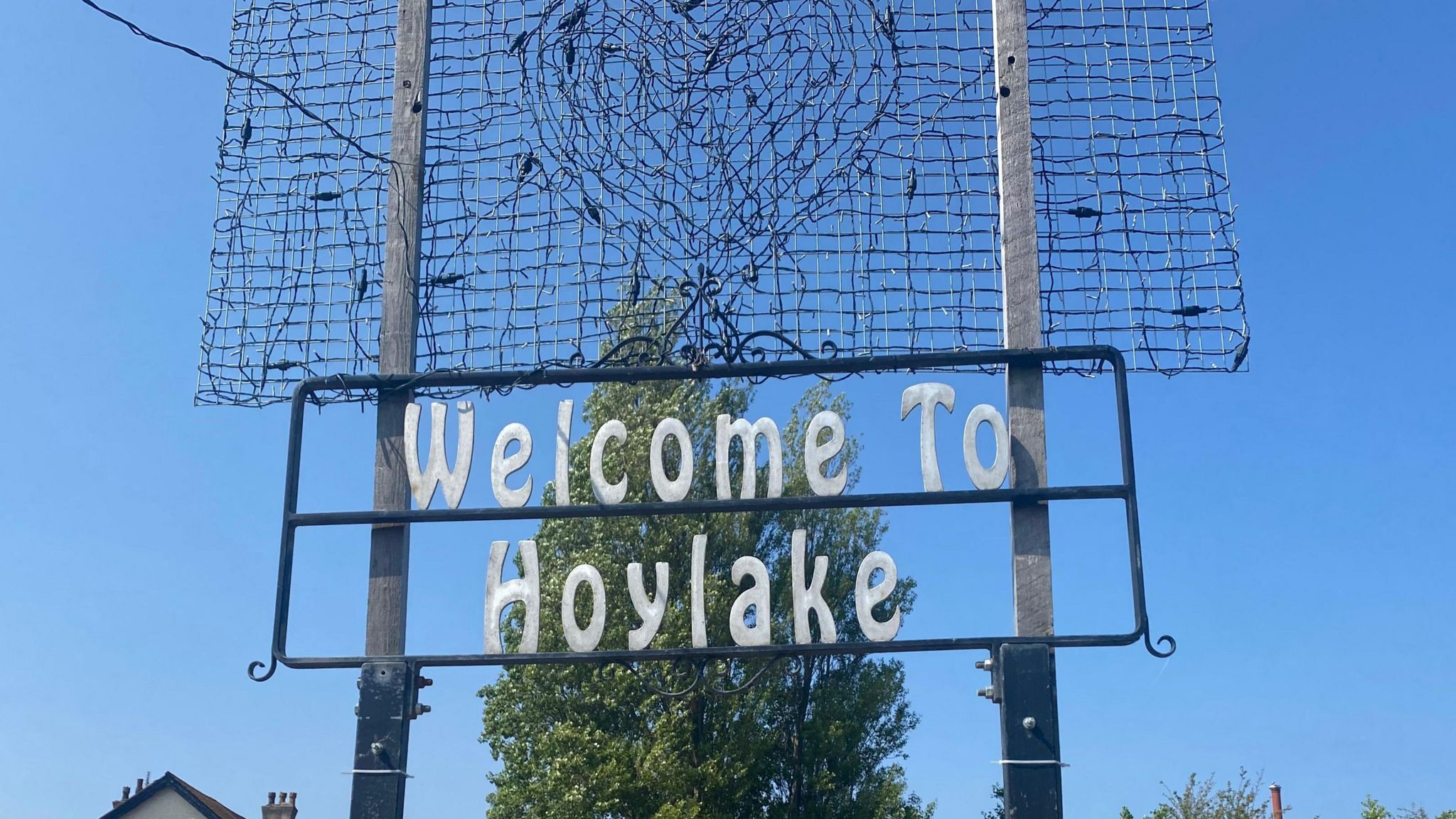A white-lettered sign reading 'Welcome to Hoylake' hangs between two grey posts, underneath an unlit heart-shaped light installation and in front of a green-leafed tree and a cloudless blue sky