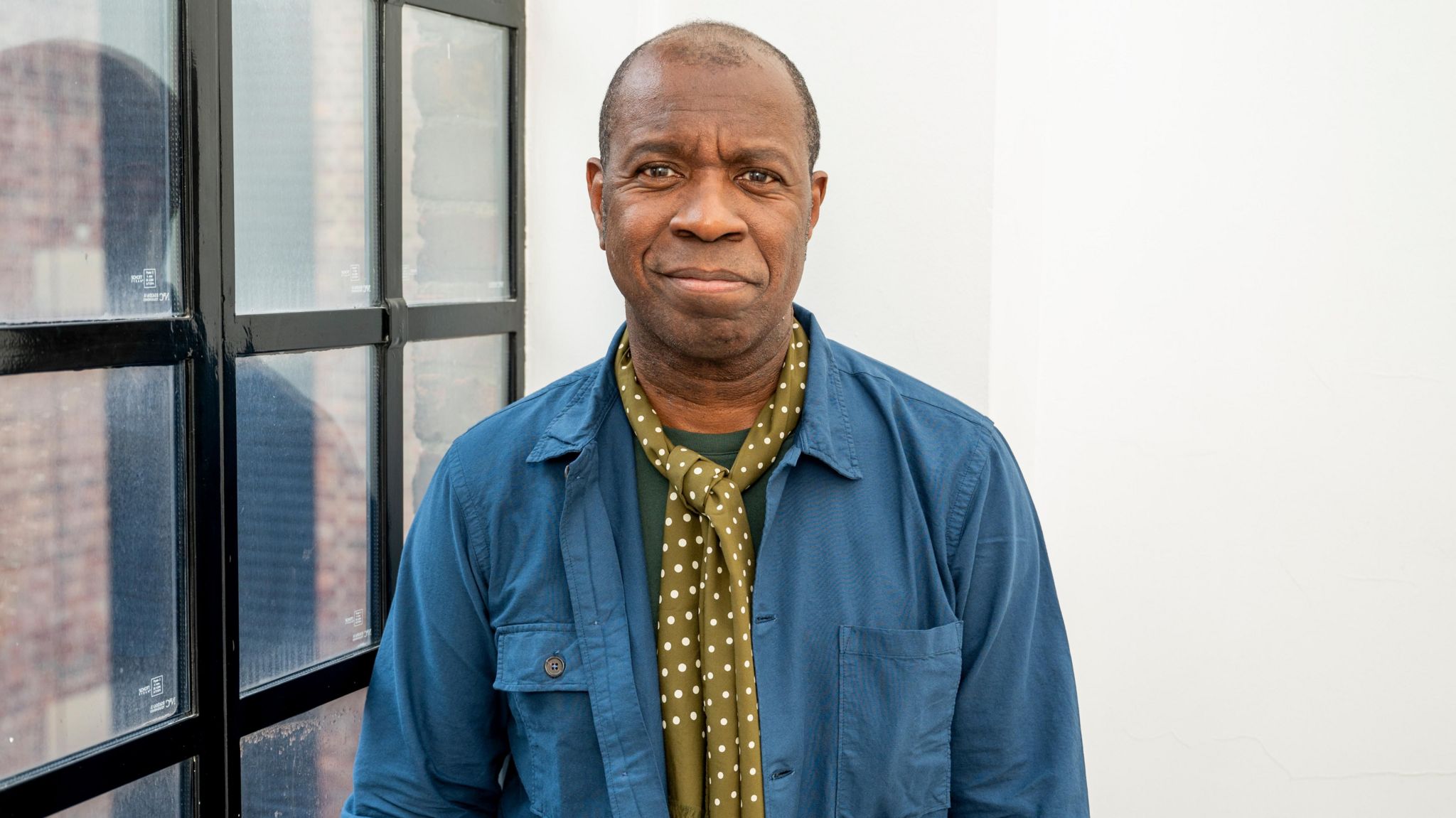Clive Myrie stood next to a white wall and window