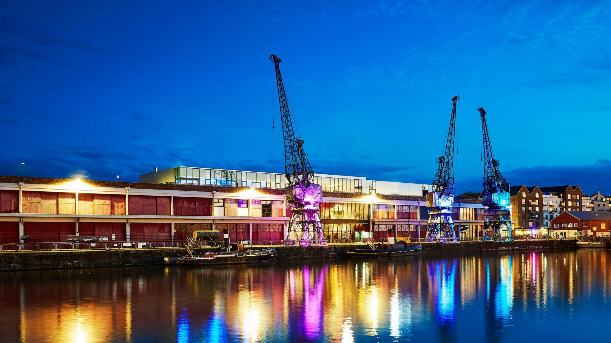 M-Shed, pictured from across the water. It's getting dark in the picture and the cranes are illuminated with purple and blue lights that reflect in the water. 