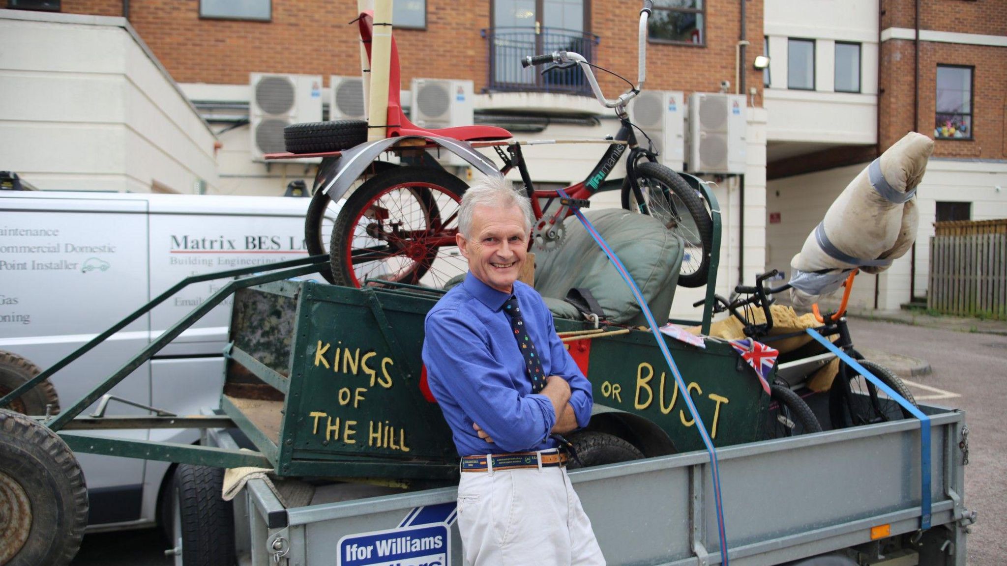 Soapbox race organiser Phil Pritchard is seen leaning against a trailer with a dark green soapbox kart inside, folding his arms.