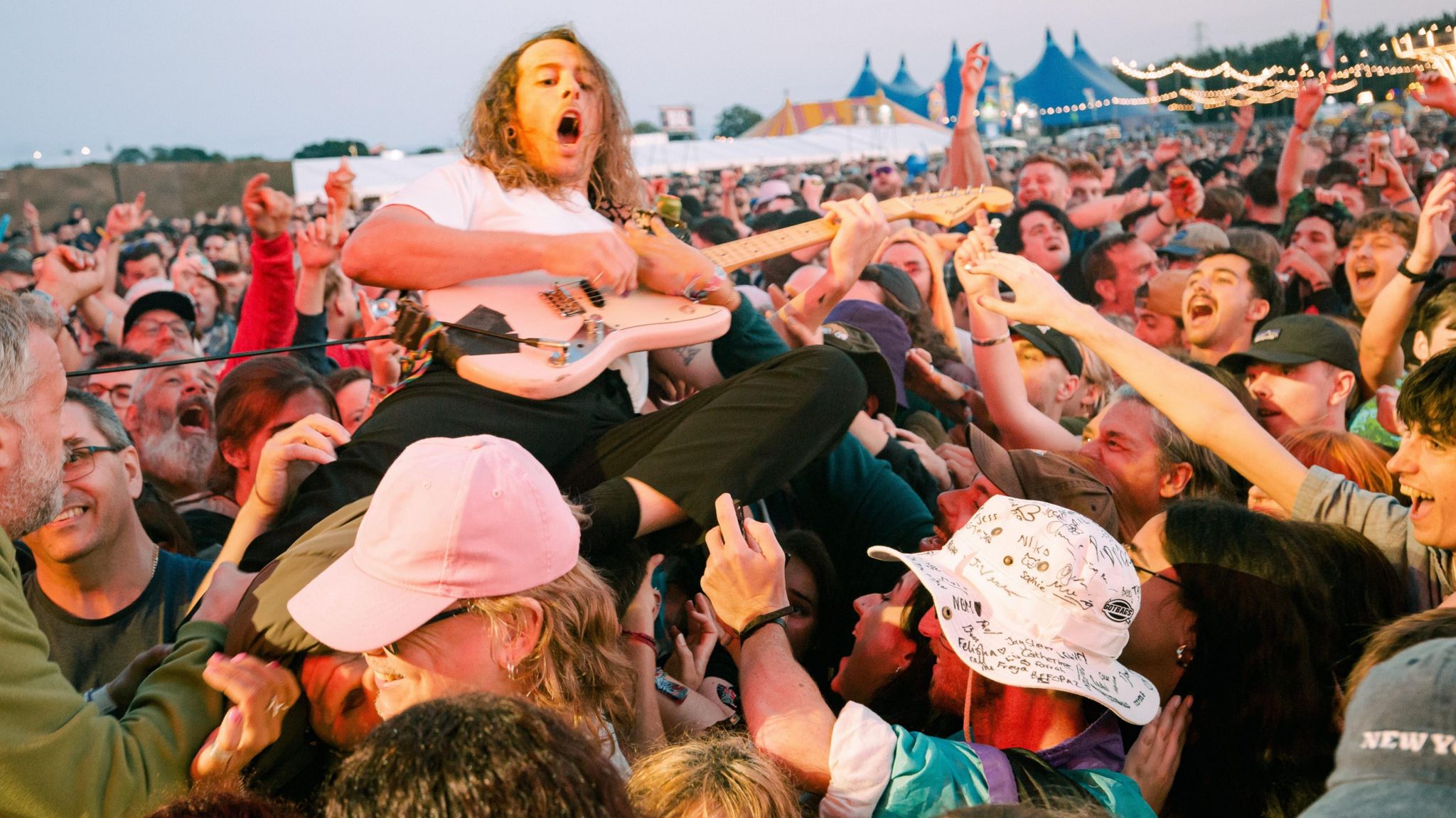 Lee Kiernan from IDLES is playing the guitar while crowdsurfing. Mr Kiernan has long dark hair and is playing a pink guitar. The crowd around him are smiling, their hands in the air.