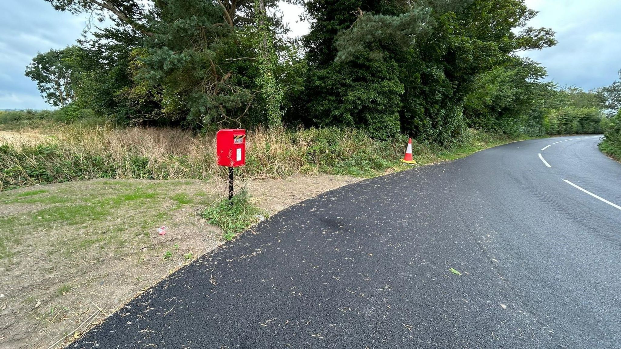 A postbox on a layby in a remote location on the sweep of a bend with the road stretching off in front.