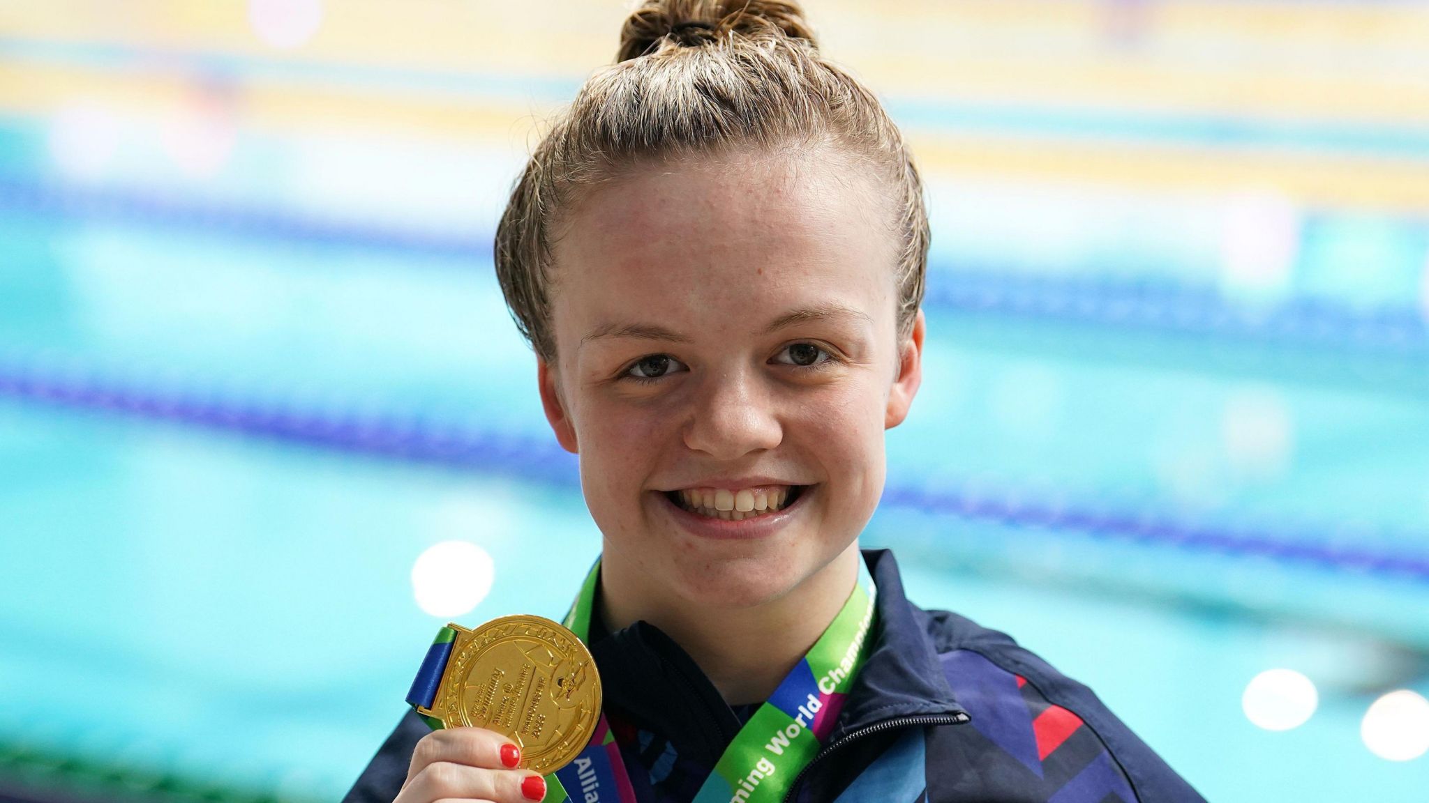 A smiling Maisie Summers-Newton stands beside a swimming pool and holds up her gold medal at the camera. 