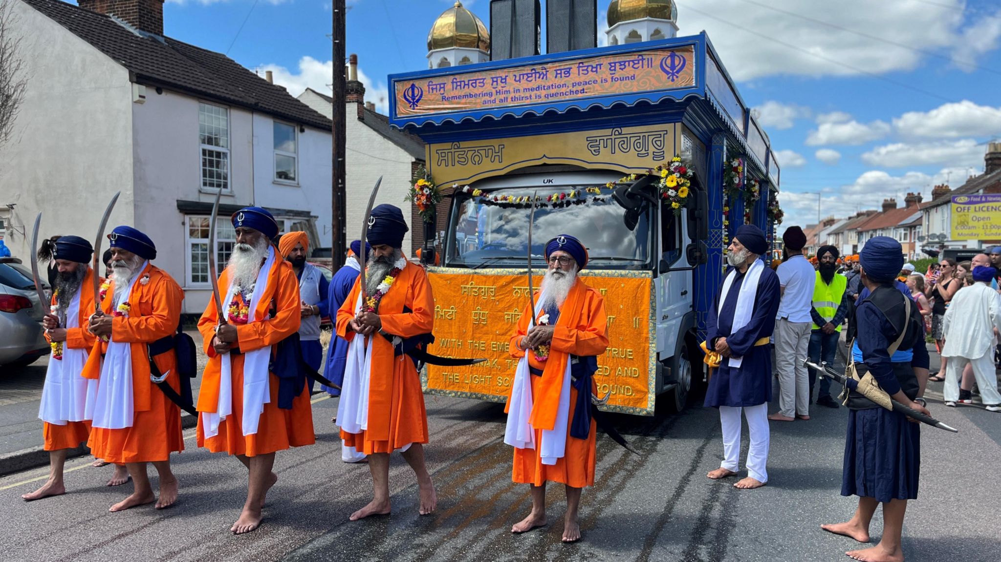 Five men in orange robes and purple turbans, carring swords in front of them, lead a procession float