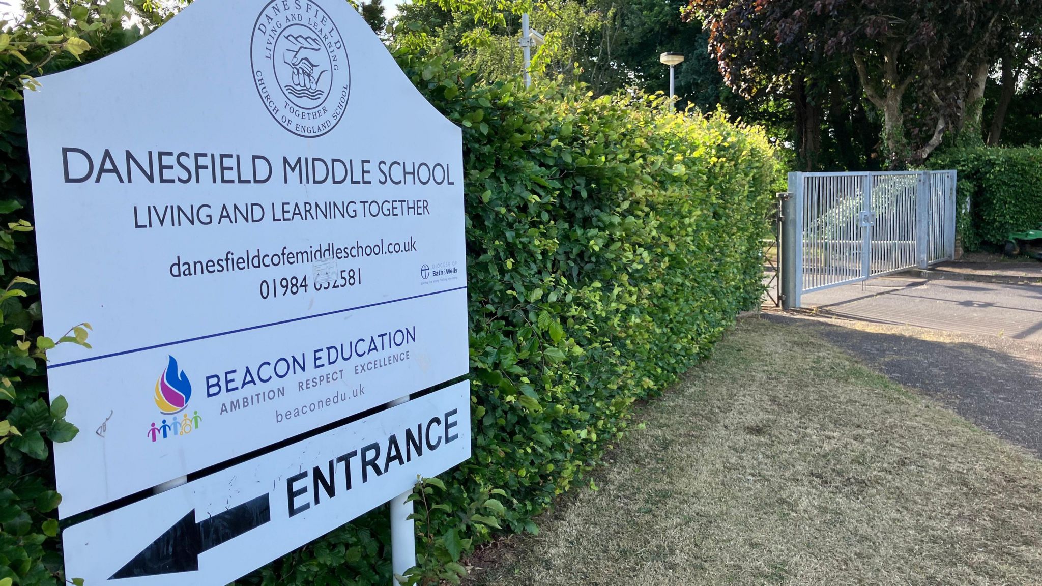 Image of the Danesfield Middle School sign. It is white, and has the logo  at the top. There is also a logo for Beacon Education. There is an arrow pointing left to the entrance at the bottom. 