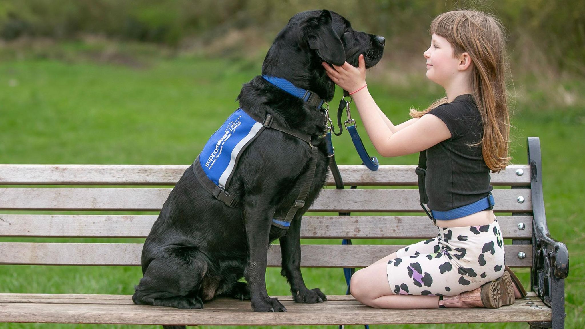 A black labrador and a young girl sitting on a park bench