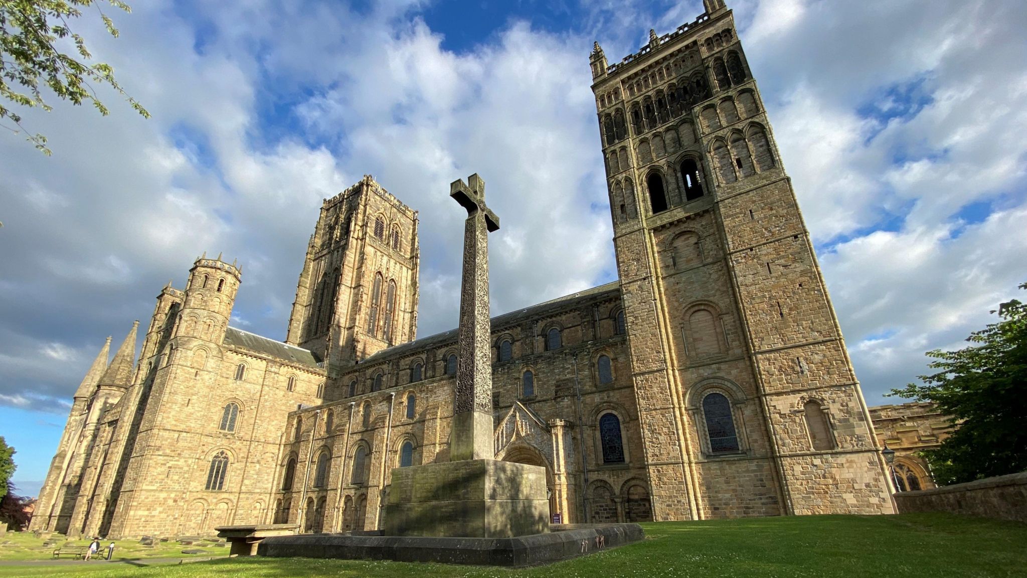 A large stone cross stands outside the front of the cathedral, a large yellow stone building with two large towers visible