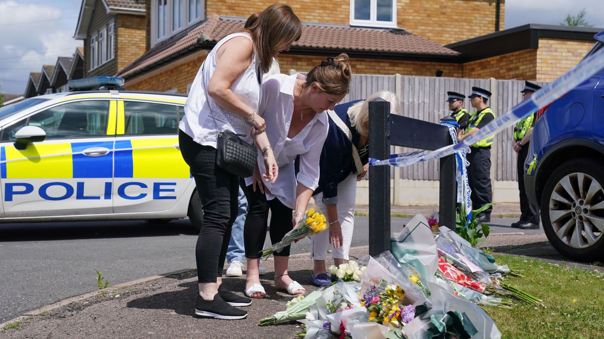 Three women lay flowers on a patch of grass at the top of Ashlyn Close, as police officers stand blocking the road