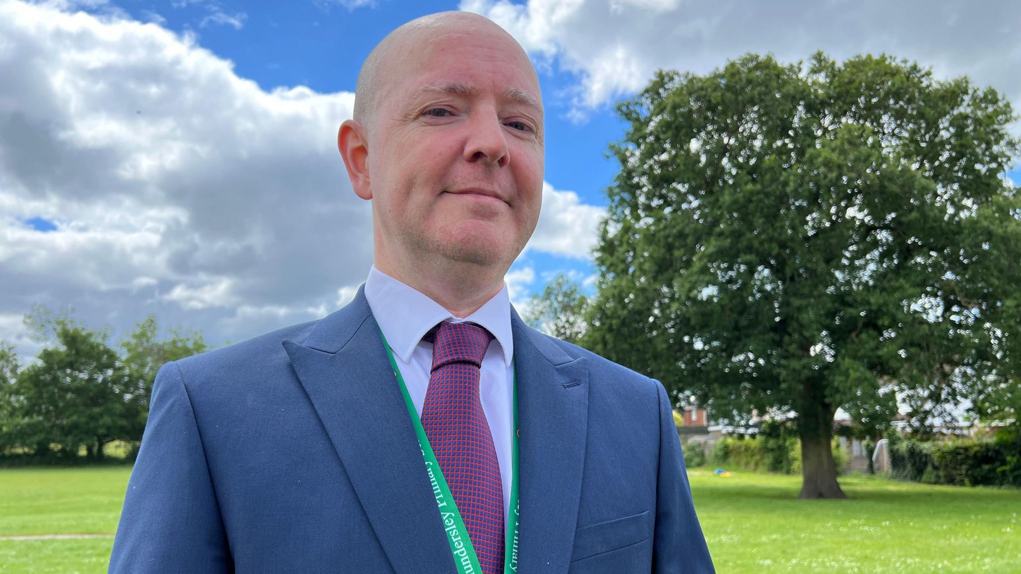 Simon English smiles and looks down at the camera dressed in a blue suit and wearing a green lanyard. Behind him is the tree in question. 