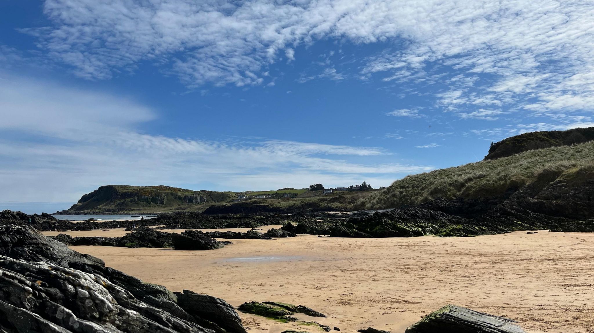 A large beach on a rocky coastline with the tide well out, and thin cloud in a blue sky above