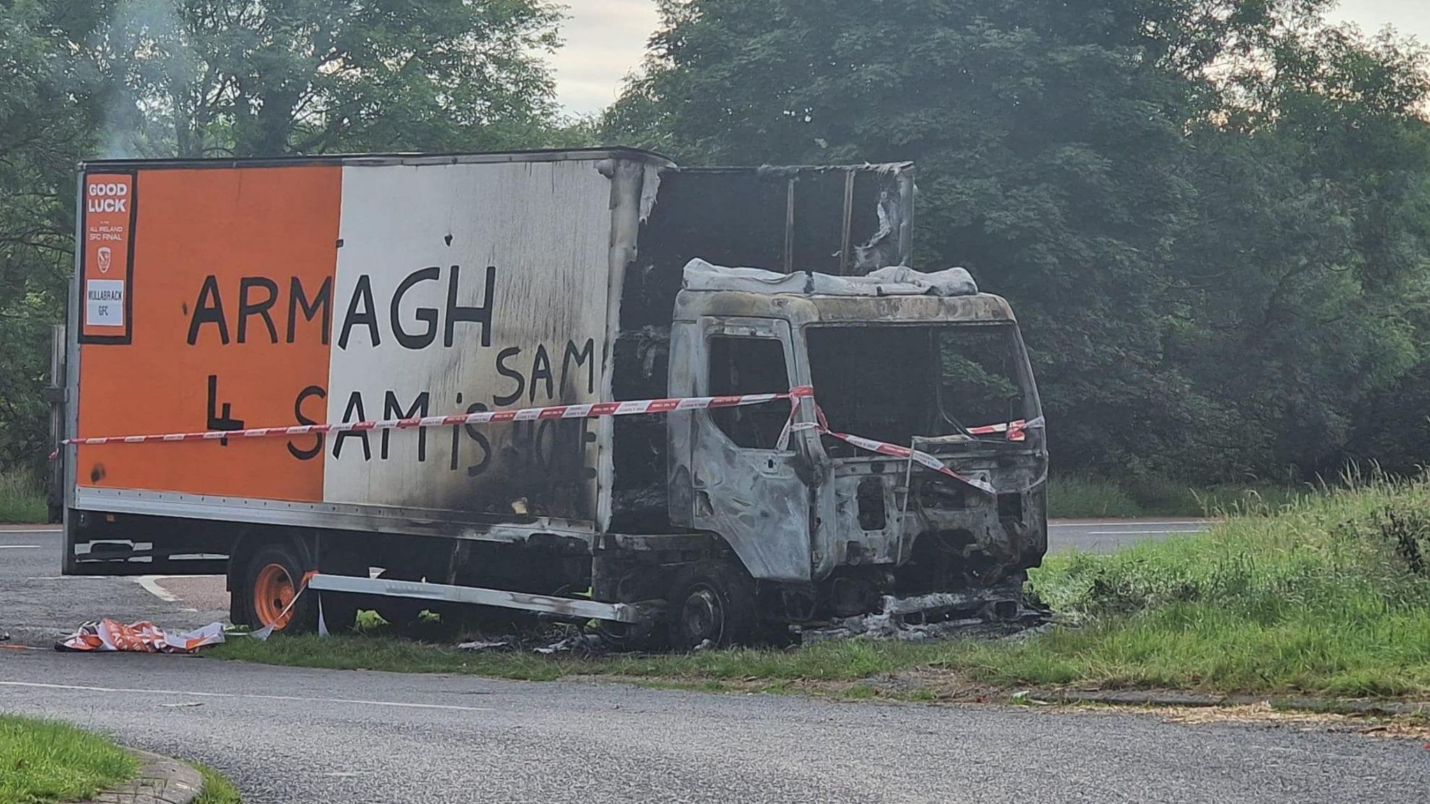 Burnt out lorry painted in orange and white with the slogan "Armagh 4 Sam"