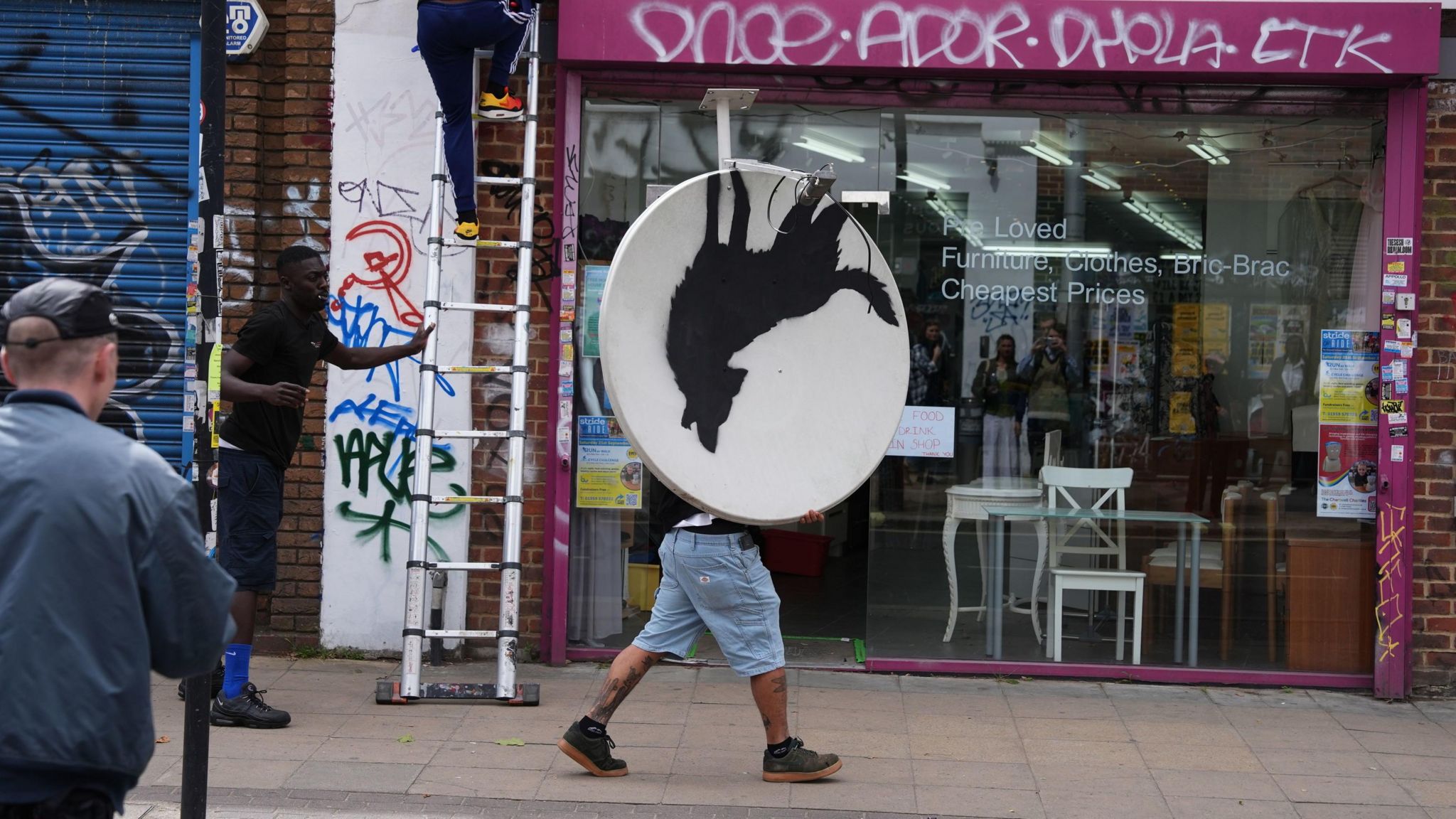 A man was seen carrying the new Banksy down the street. His top half is obscured by the satellite dish which is being carried so the artwork is upside down. He is wearing light blue shorts and dark shoes and is walking in profile along the pavement.