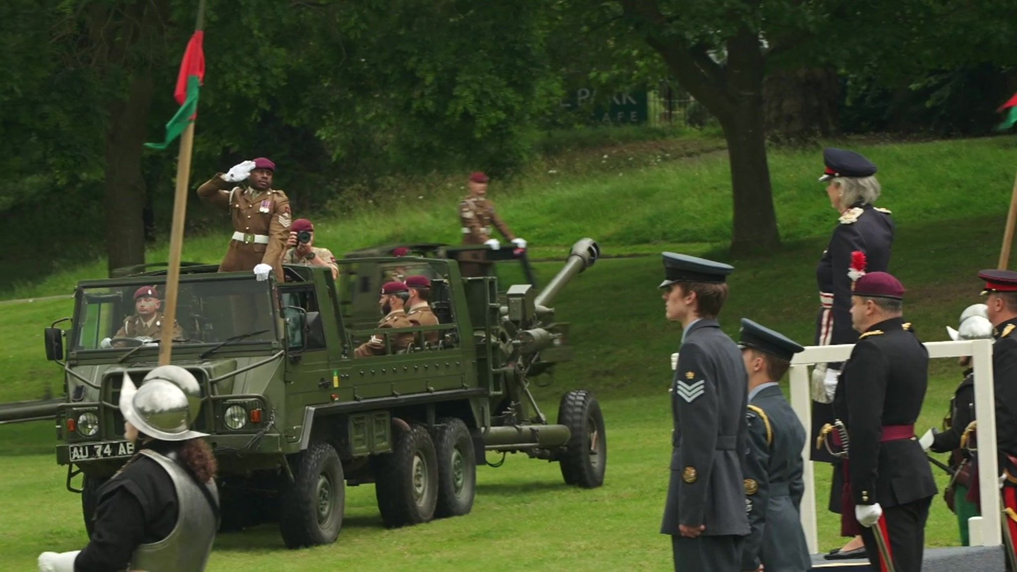 Two army vehicles, one with a soldier saluting, pass by the saluting dais