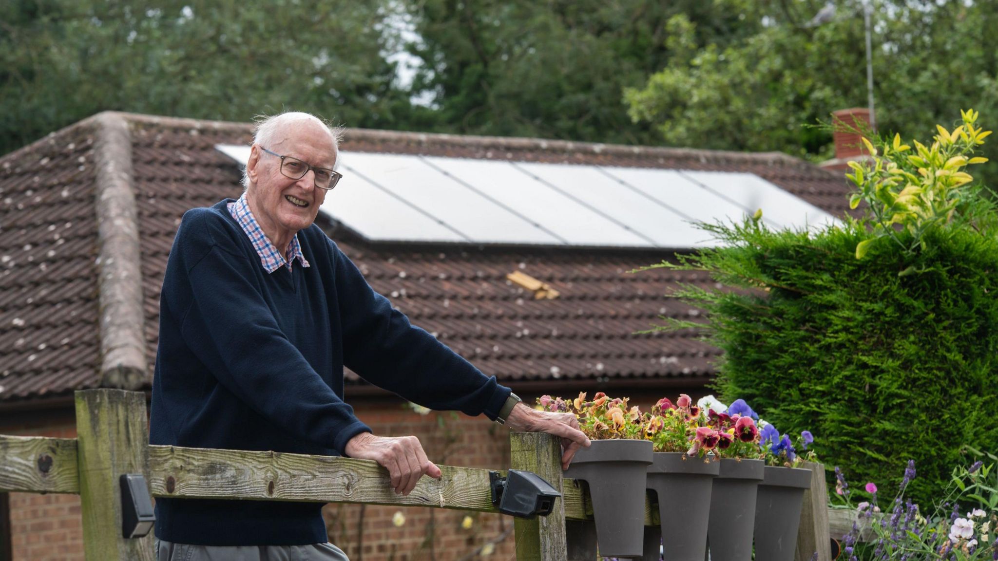 Geoffrey Evans stands in front of a wooden fence, wearing a chequered shirt with a navy jumper. He has glasses, white hair and is balding. A row of flower pots are fixed to the fence on his left, a brick house with solar panels on the roof is in the background.