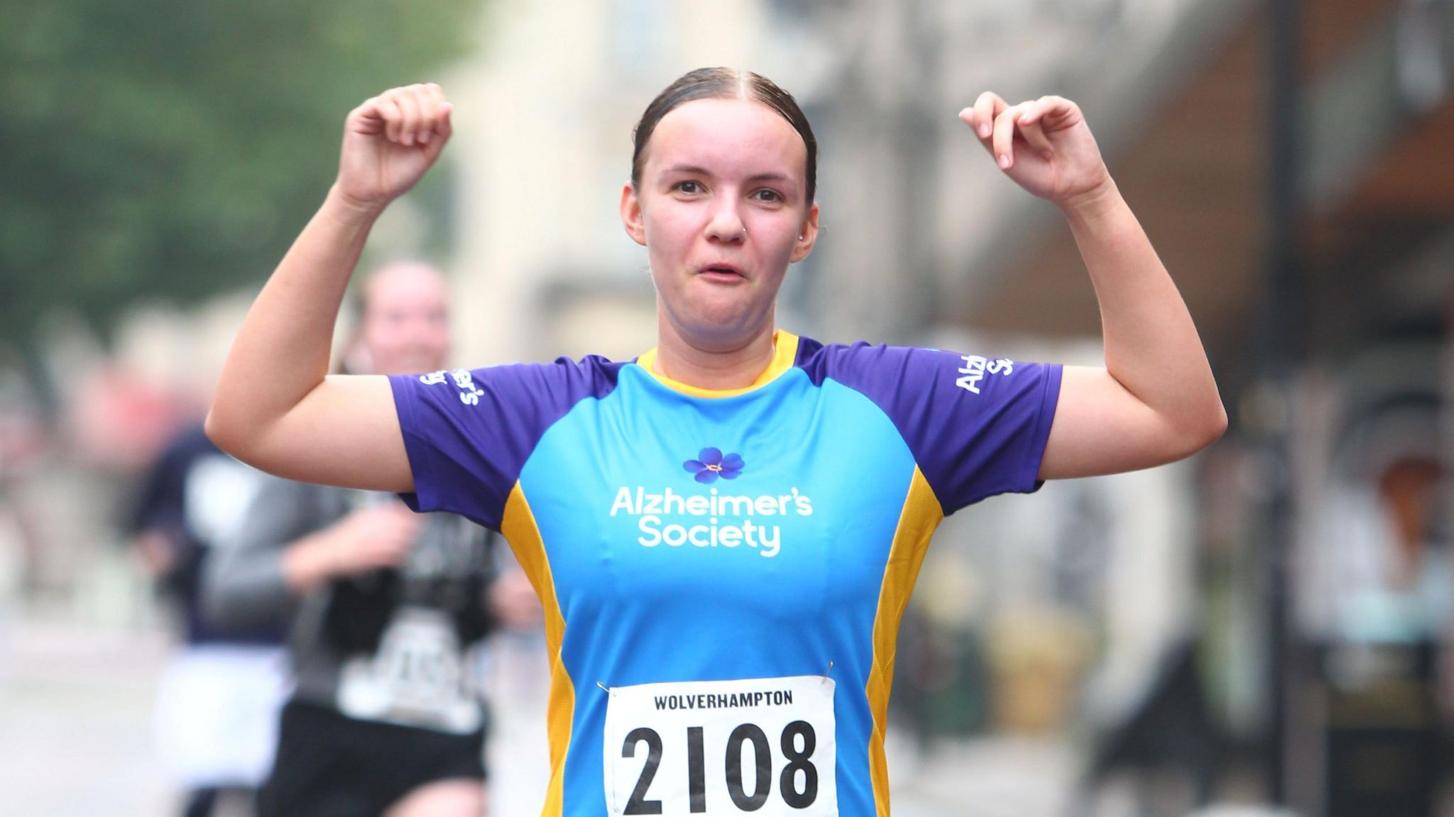 A woman in a blue top raises her arms in celebration as she looks at the camera