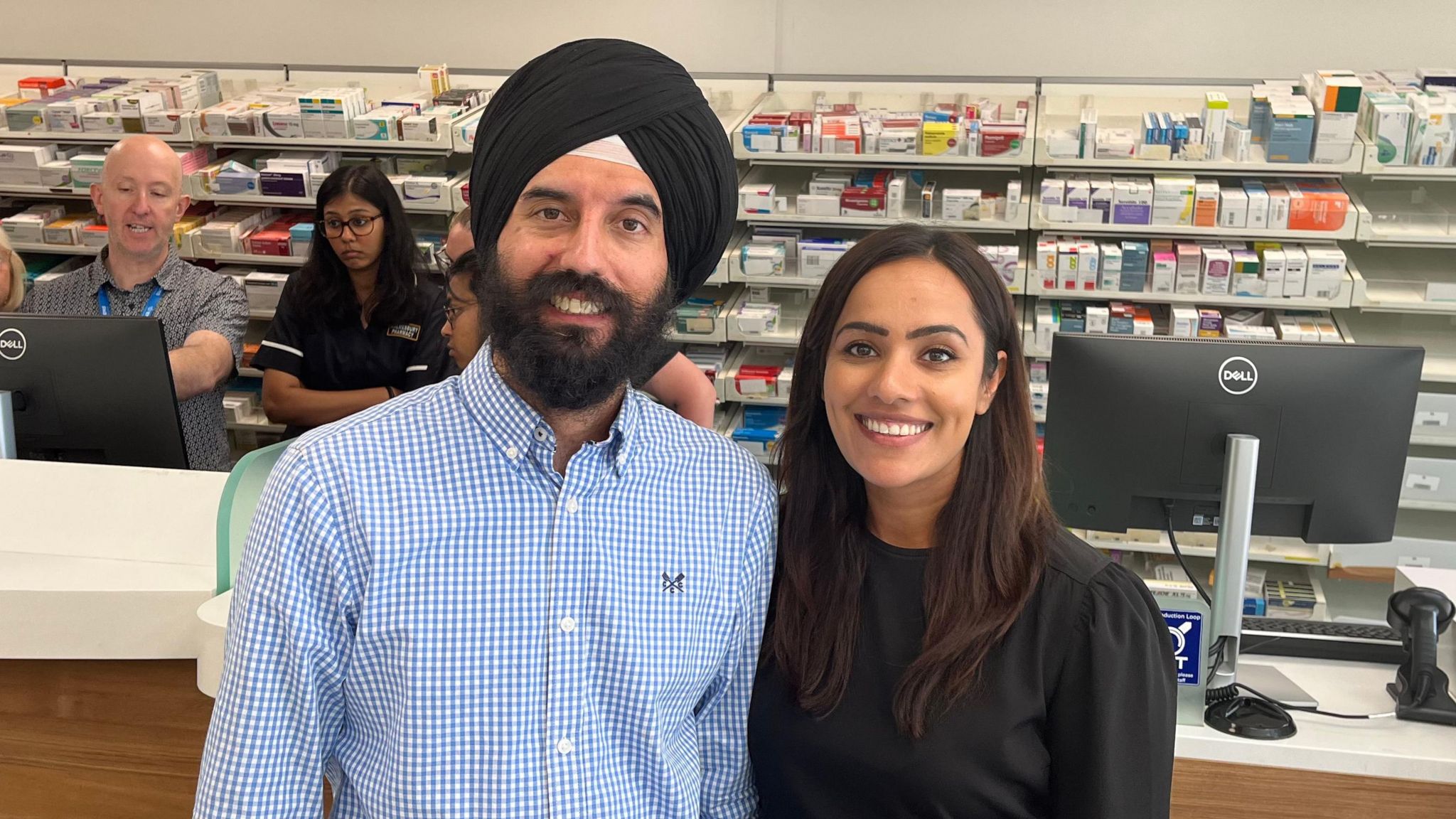 A man wearing a blue shirt and black turban standing and smiling with a woman inside a pharmacy in Malmesbury. Customers are visible in the background