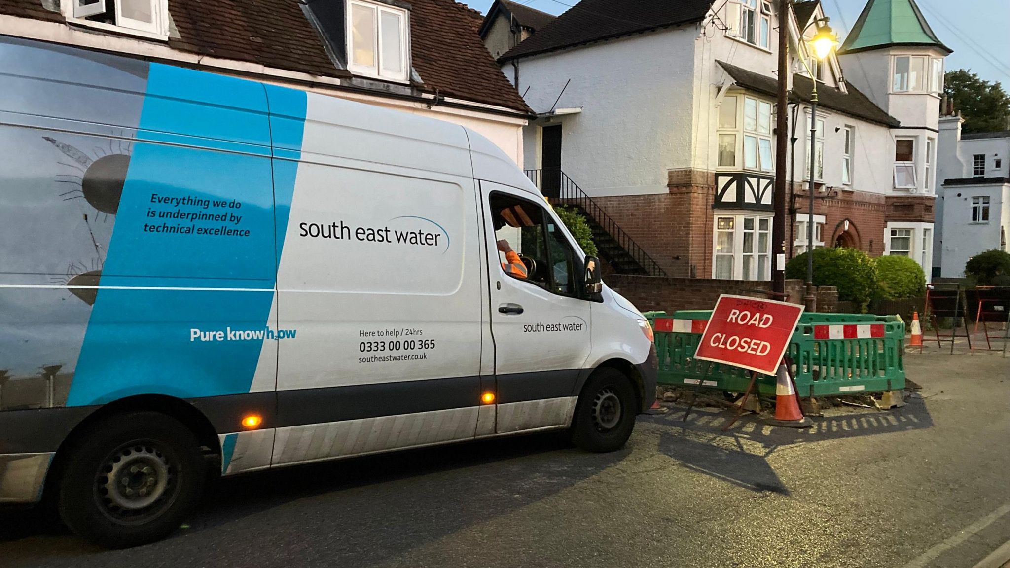 A white transit van is parked in a residential street with a road closure sign in front.