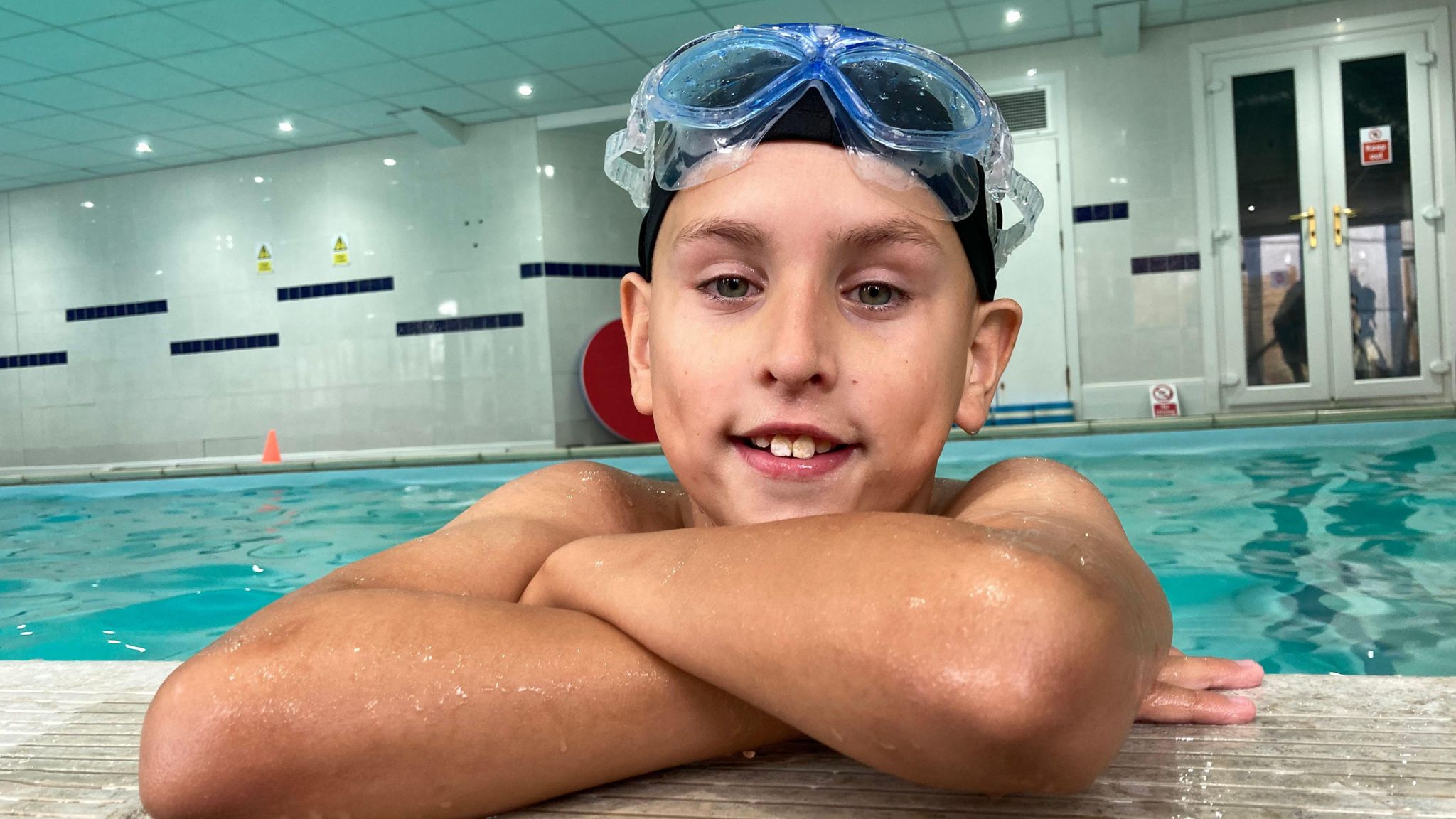 Jayden in a pool, leaning on the side, with goggles and a swim cap on his head.