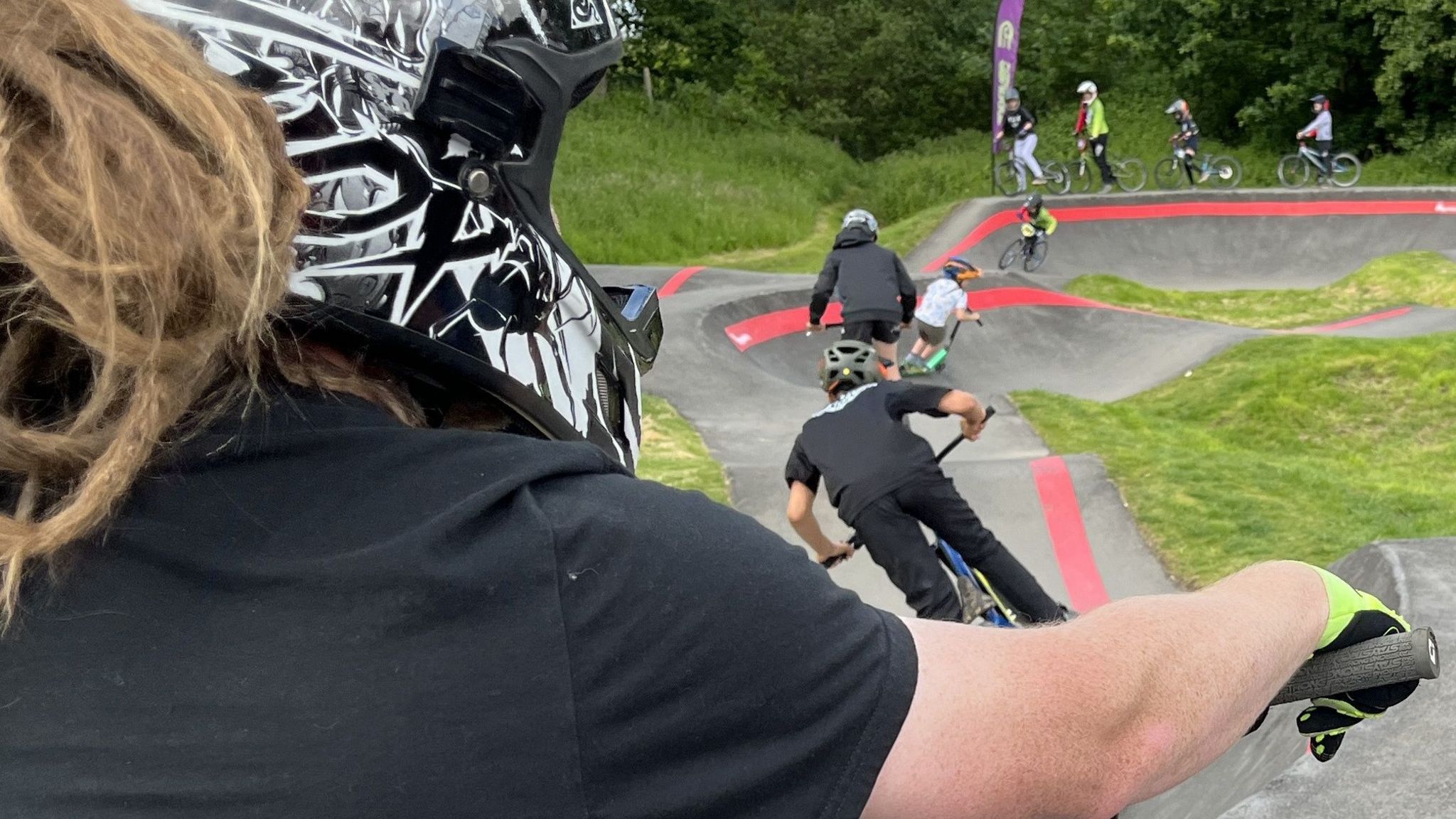Profile shot of a BMX rider with dreadlocks showing at the back of their crash helmet with riders of all ages on the track in front