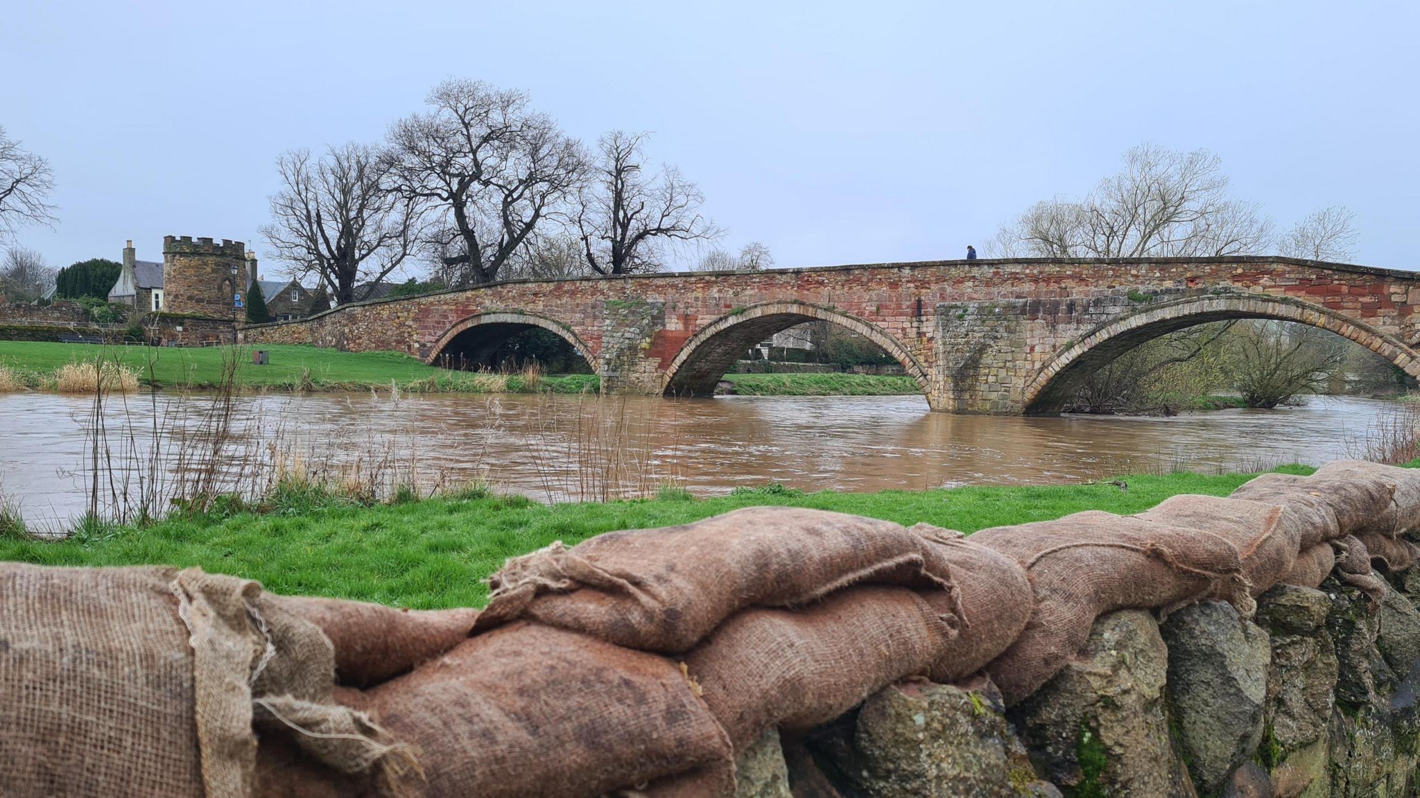 Photograph of a swollen river that has filled out onto the flood plain.  In the foreground a stone wall that has sandbags laid on top.