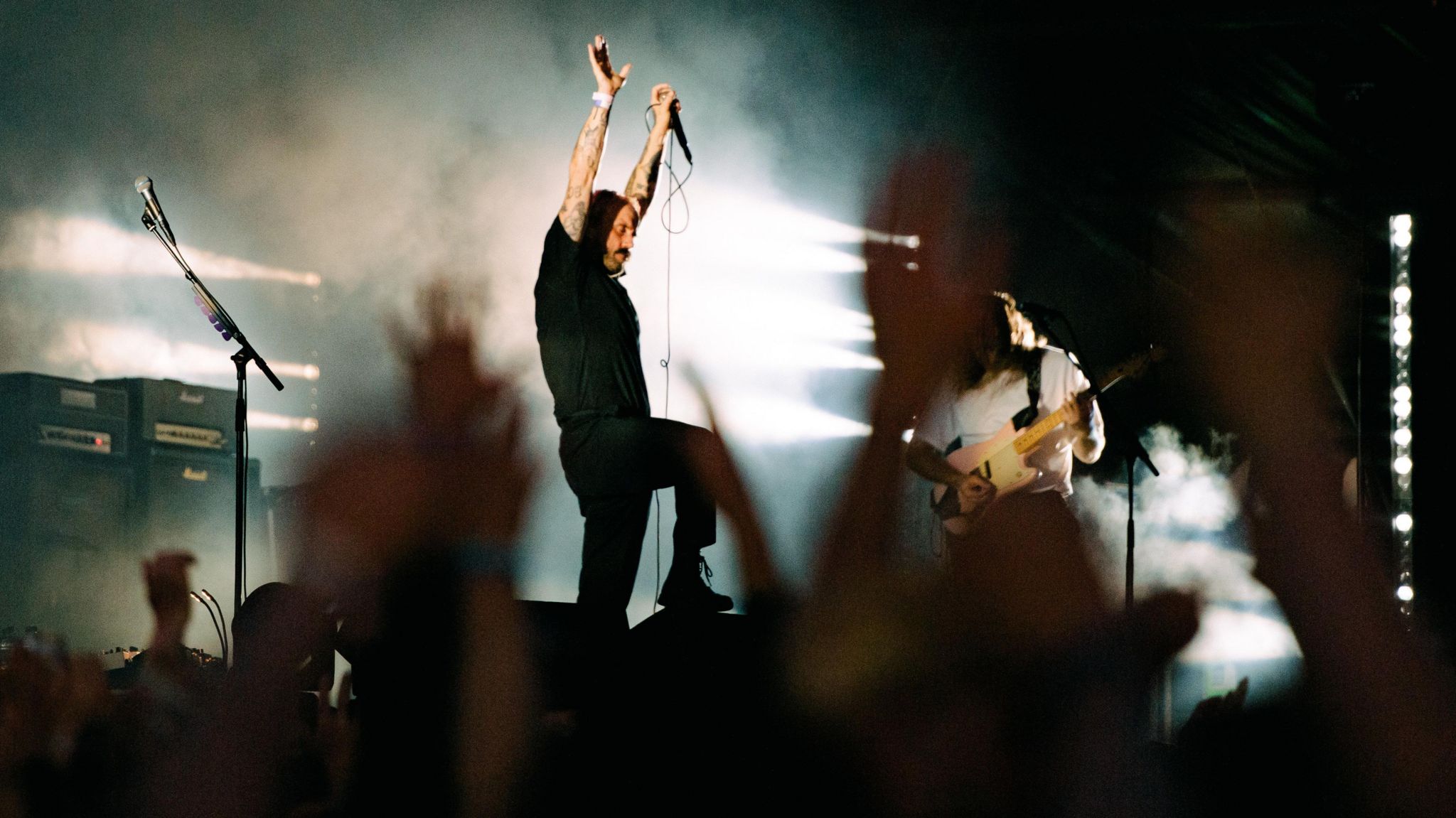 Joe Talbot, the lead singer of IDLES, has his hands aloft on stage, one foot perched on a speaker. Hands are in the air in the foreground.