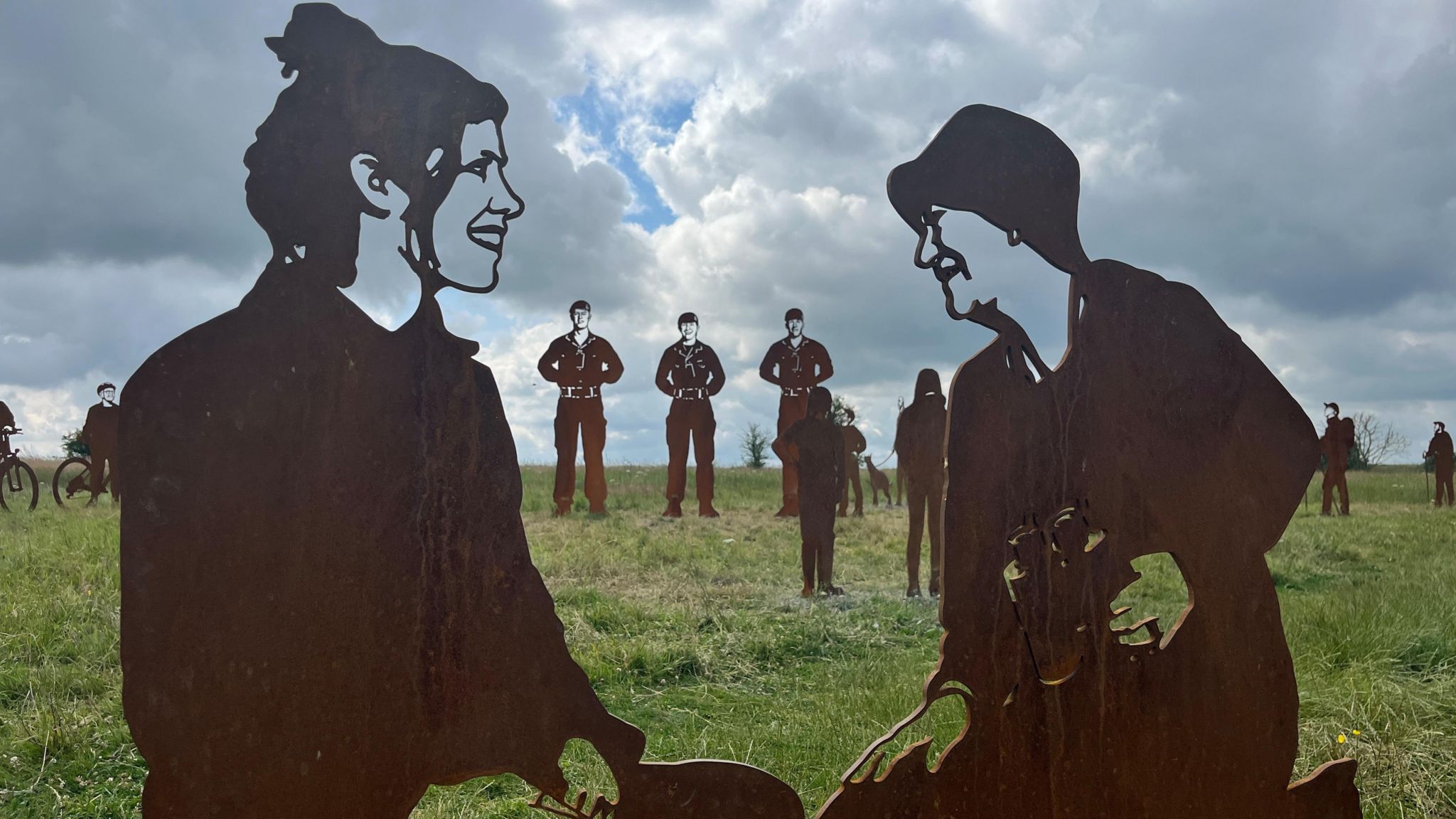 Metal statues of a mother and daughter in a field on Salisbury Plain, with further statues of three soldiers in the background