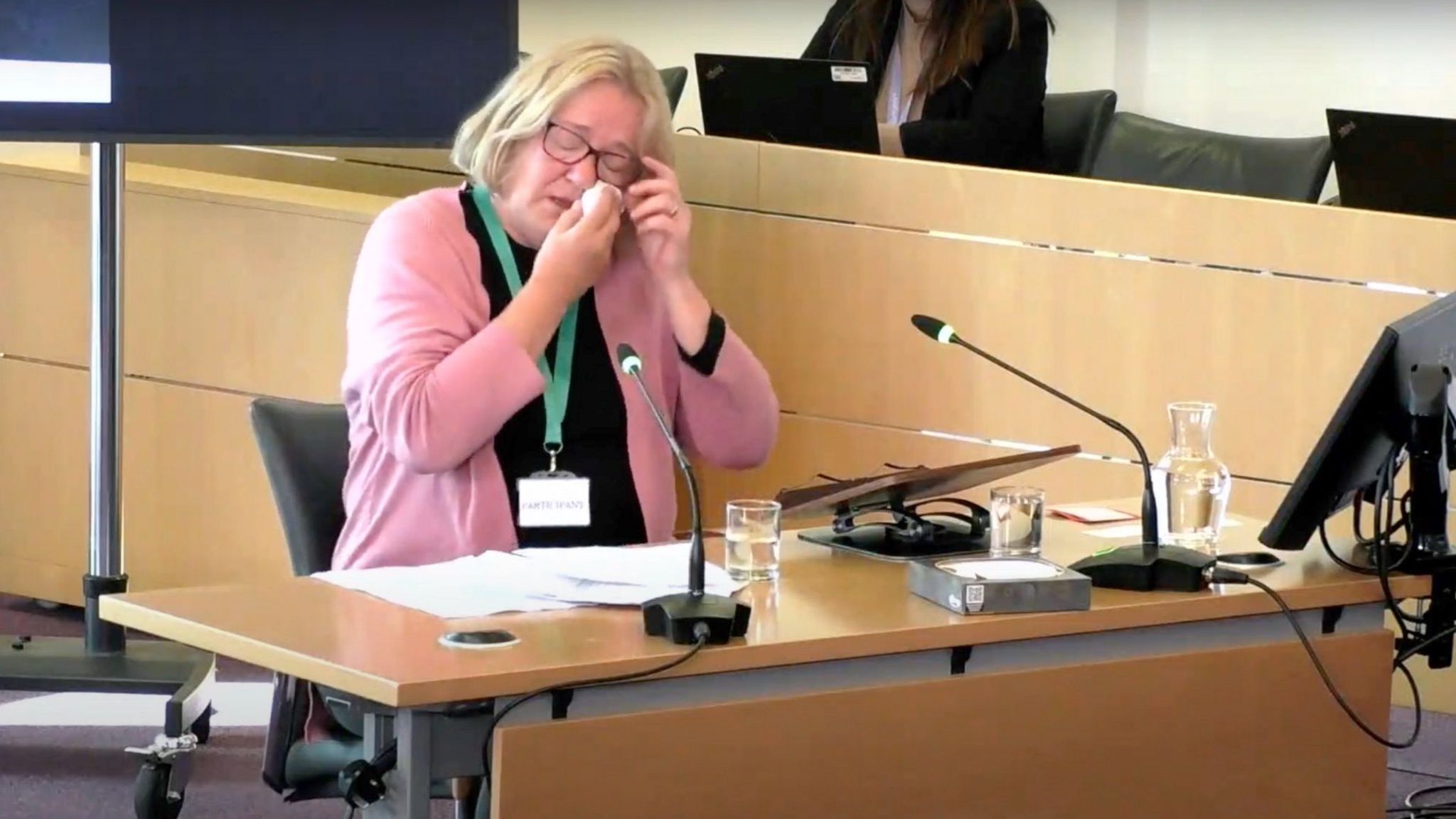 Karon Pimm sitting behind a desk at the inquiry, raising her glasses slighty and wiping her eyes. She is wearing a pink cardigan.