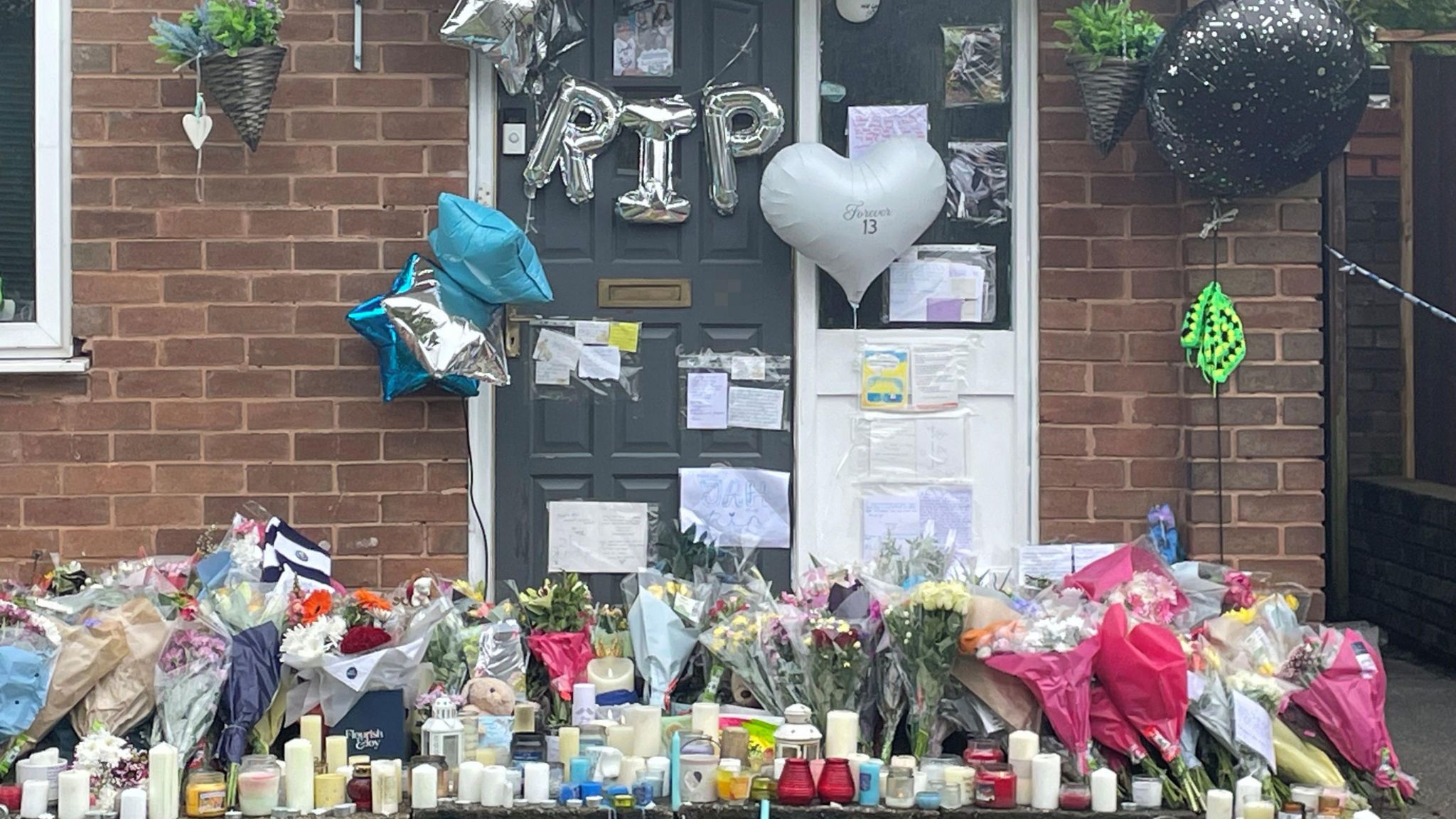 Candles, floral tributes and balloons outside a house. There are silver balloons spelling out the letters "RIP" hanging across the house's front door.