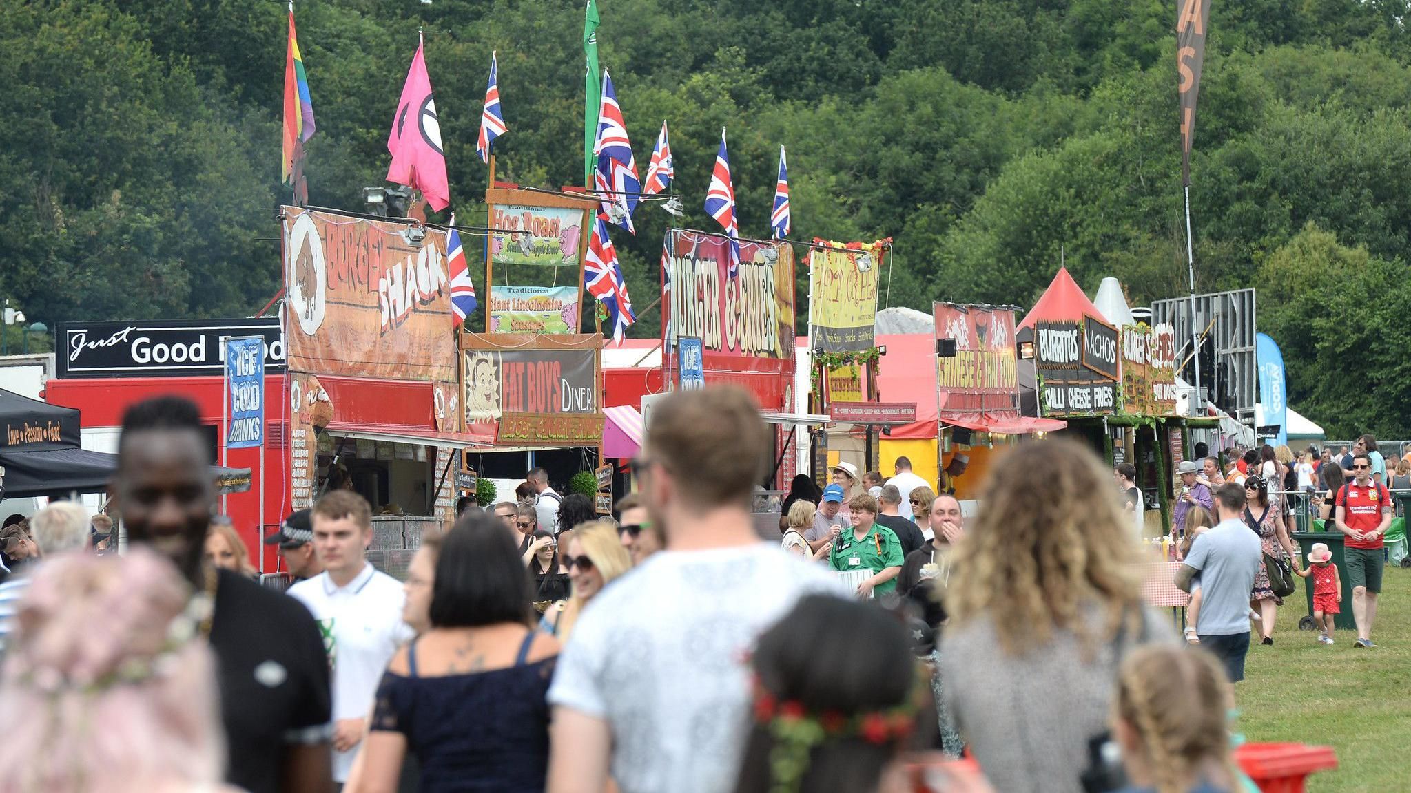 The backs of festival goers at the Godiva Festival. In front of them are a range of stalls with many having flags on top of their wooden signs