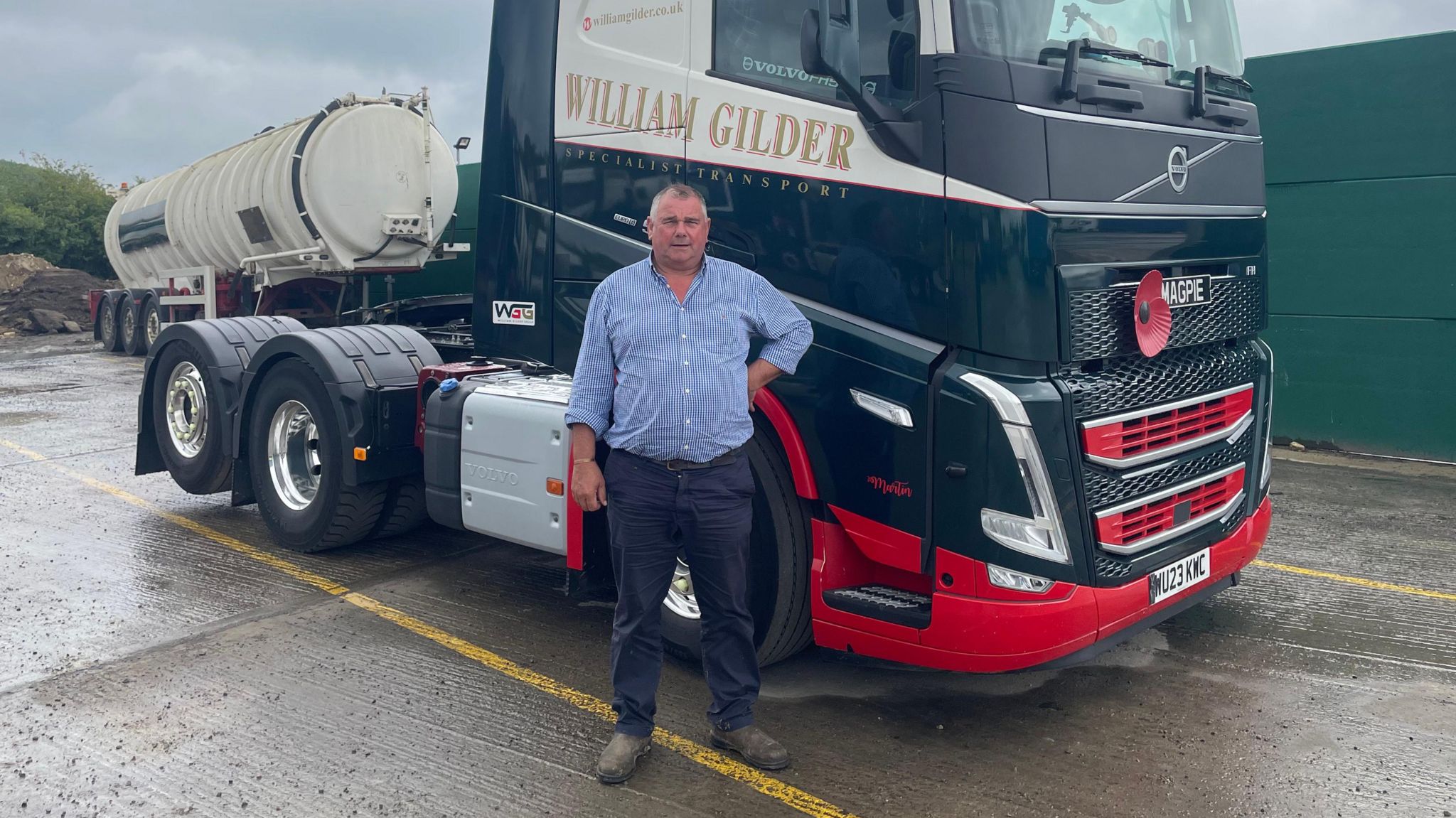 William Gilder stood by one of his lorries. He is wearing a shirt and trousers and has his hand on his hip. 