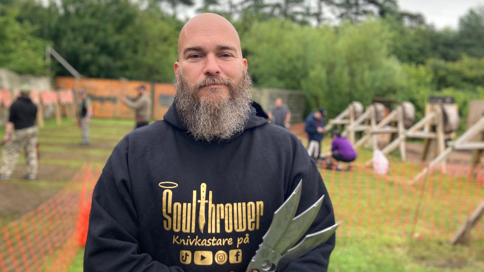 Marcus Pehart is shaven-headed with a bushy grey beard. He wears a black hooded top with gold letters reading "Soulthrower" and holders three knives. In the background is a green grassy area where people are throwing knives towards wooden targets.