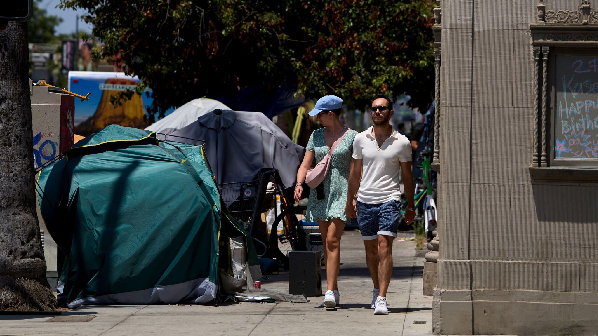 A man and woman walk on a sidewalk in Los Angeles as the woman peers over at several tents where homeless people are residing. 