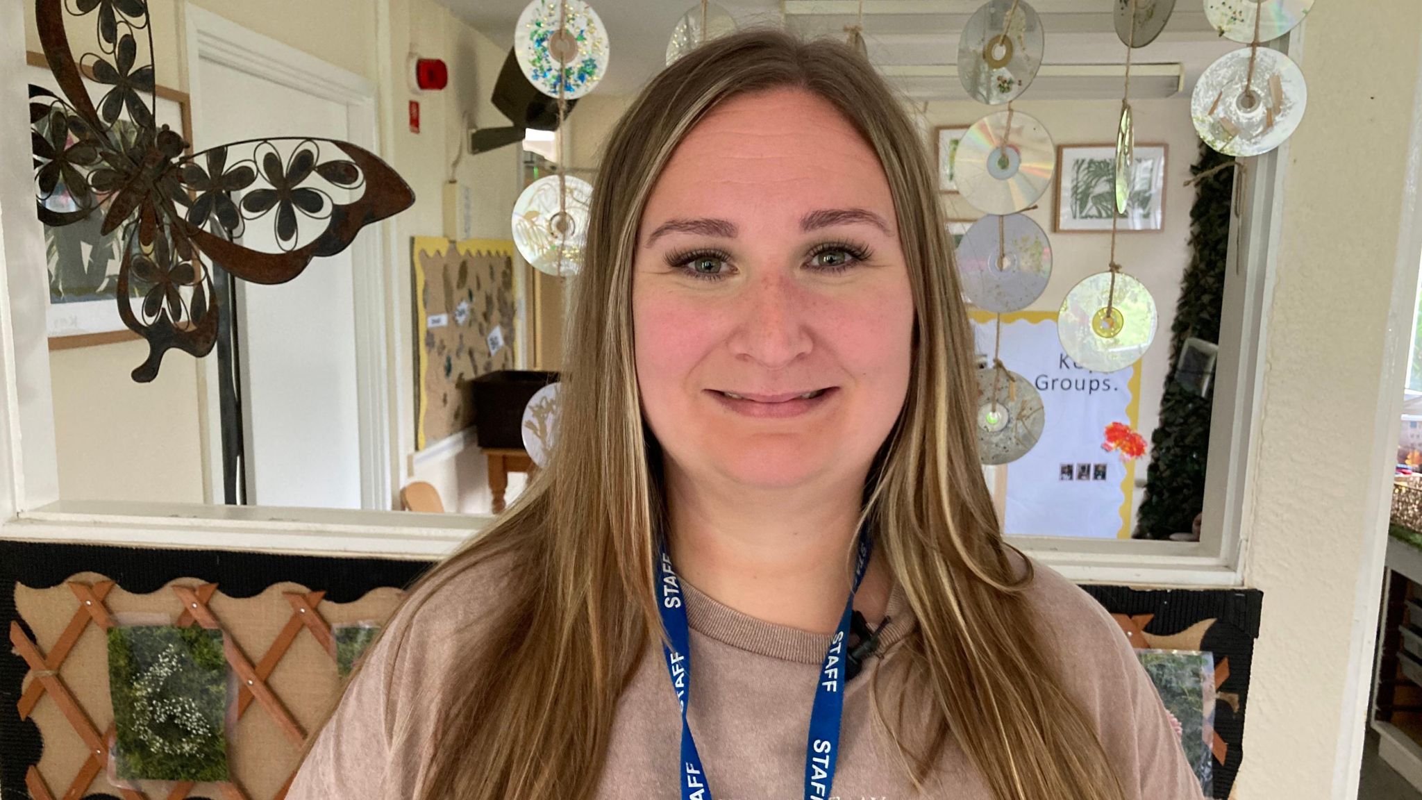 Rachel Britton smiles at the camera in the nursery. She has long dark blonde hair and wears a blue lanyard with "staff" written on it. 