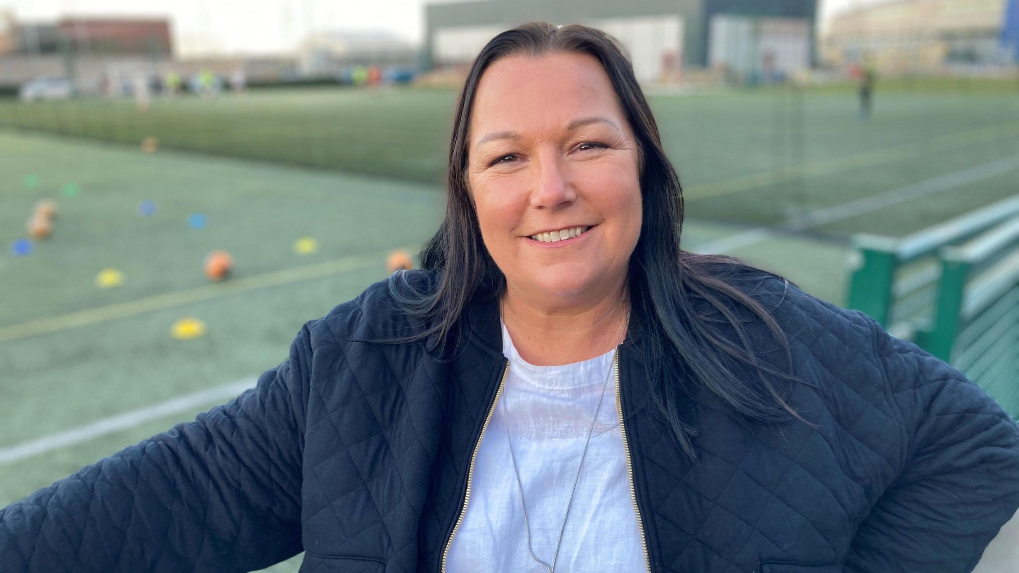 Alison McGee smiling at the camera. She is wearing a white shirt and a black jacket. She has long straight black hair. The football pitch is behind her. 