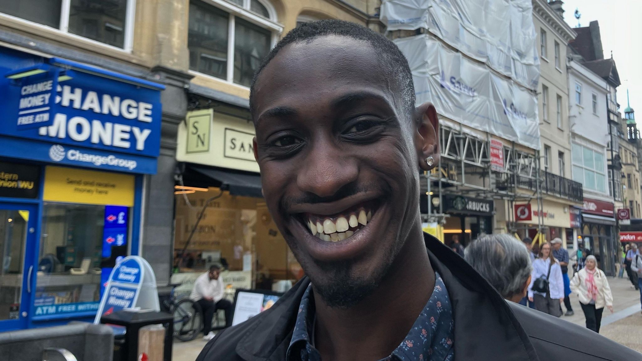 Samuel Ouyewusi is black and is smiling for the camera. He has a diamond earring in his left ear and is wearing a blue patterned shirt with a black coat 